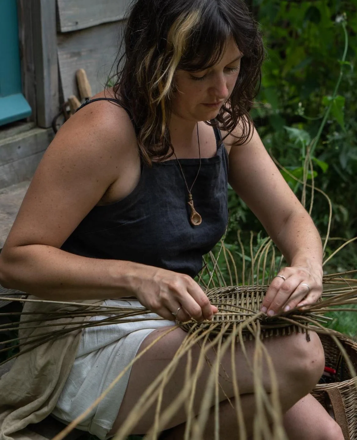 A woman with dark hair and a natural-haired blonde streak, wearing a black sleeveless top and cream shorts, is weaving a basket outdoors surrounded by greenery.