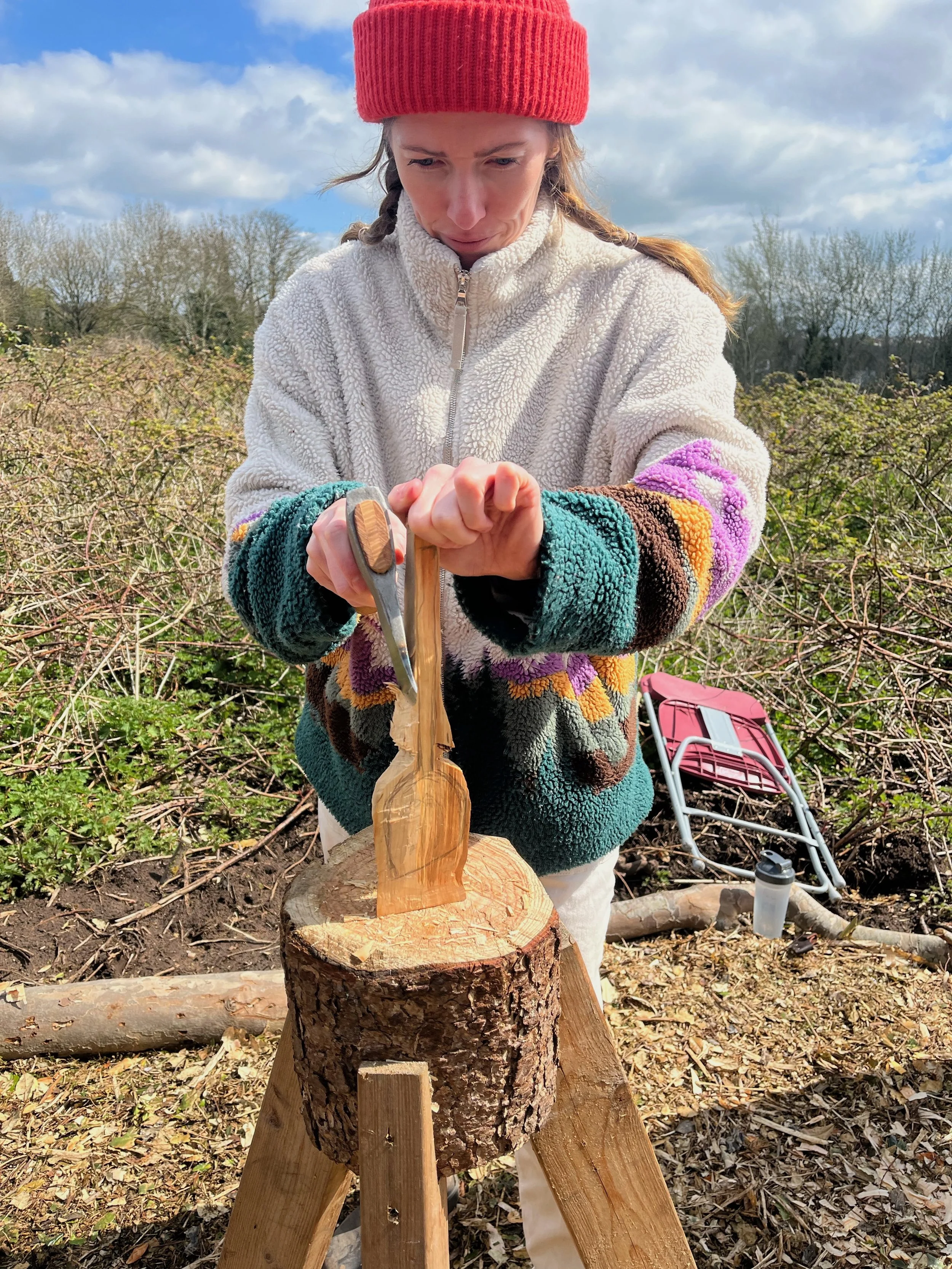 A woman in a red beanie and colorful fleece jacket is carving a figure into a piece of wood using a carving gouge, outdoors in a rustic setting with bushes, trees, and a blue sky with clouds.