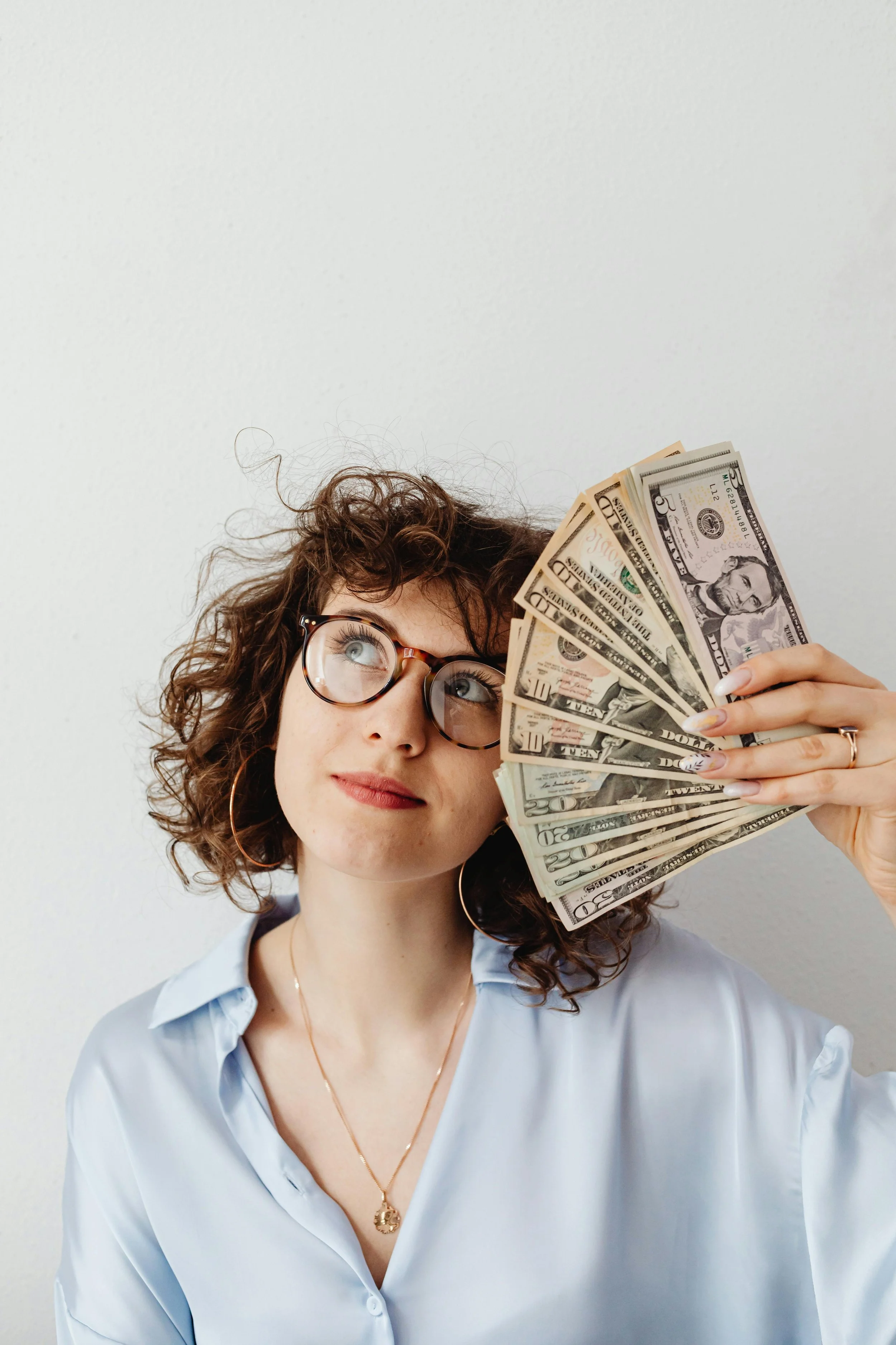 Une femme avec des lunettes, des boucles de cheveux, portant une chemise bleue, tient des billets de banque américains en faisant semblant de les regarder.