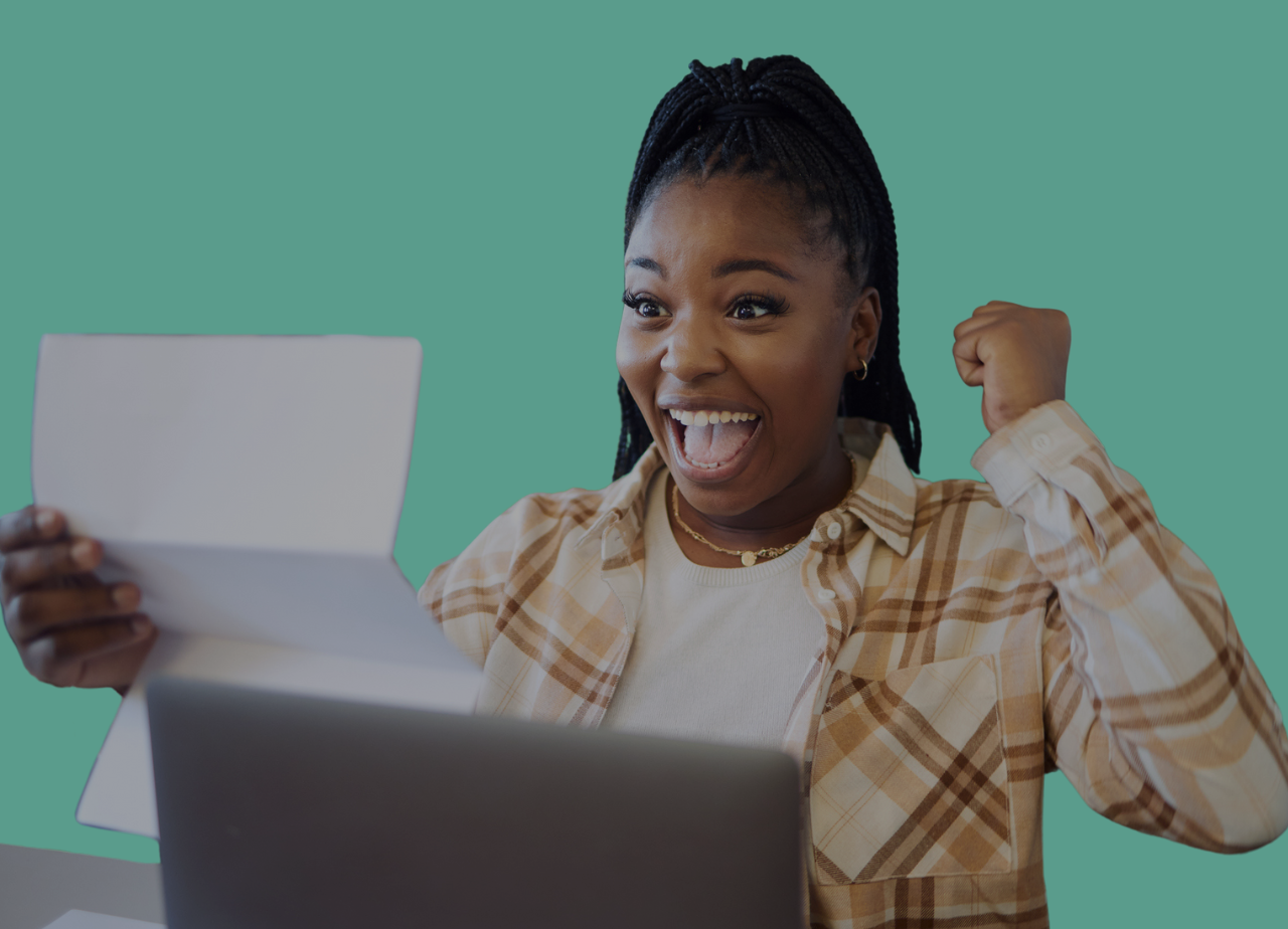 Une femme afro-américaine souriante, levant le bras en signe de victoire, regarde un ordinateur portable et tient une tablette. Elle exprime la joie ou la réussite.