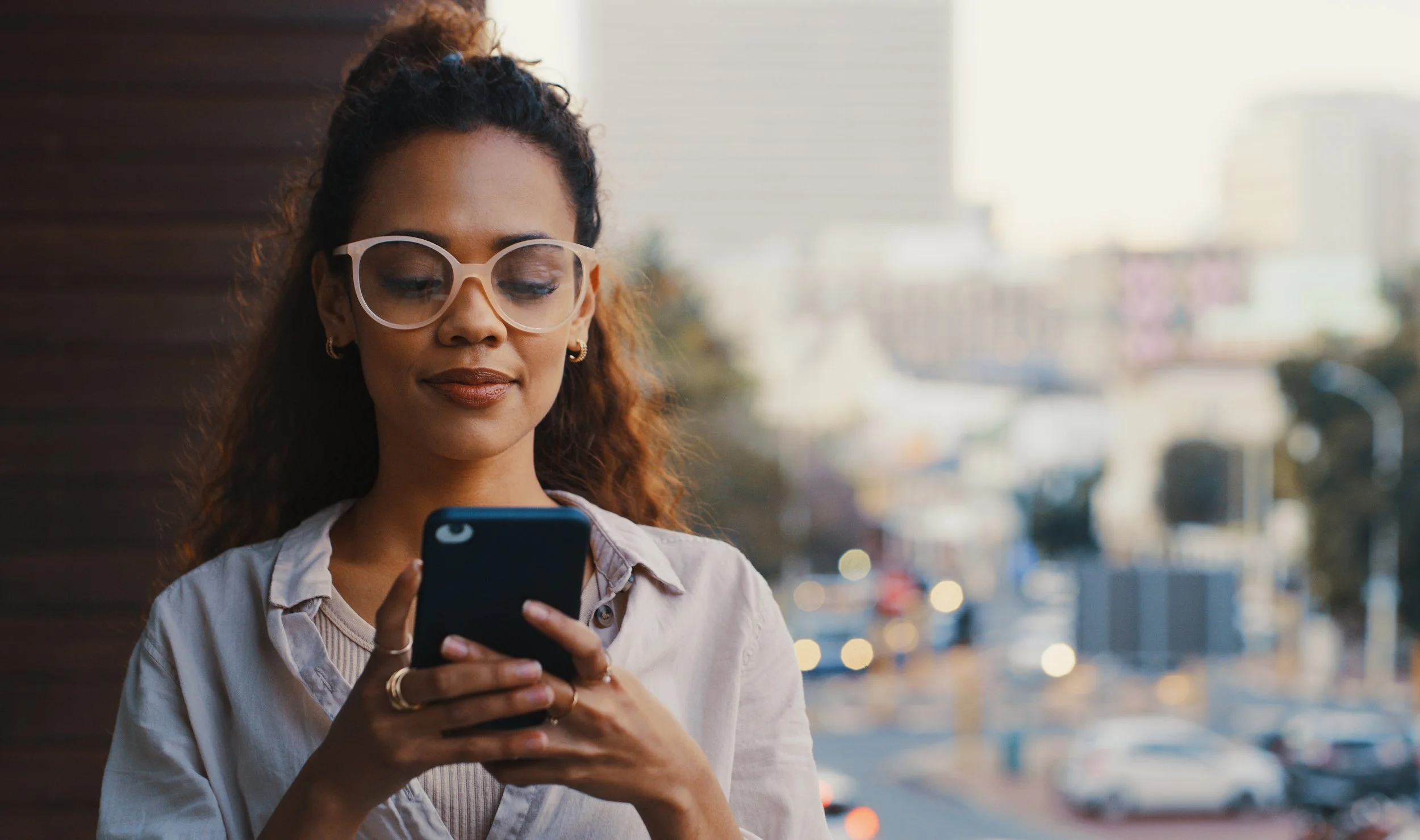 Jeune femme portant des lunettes, tenant un smartphone, en arrière-plan un paysage urbain flou.