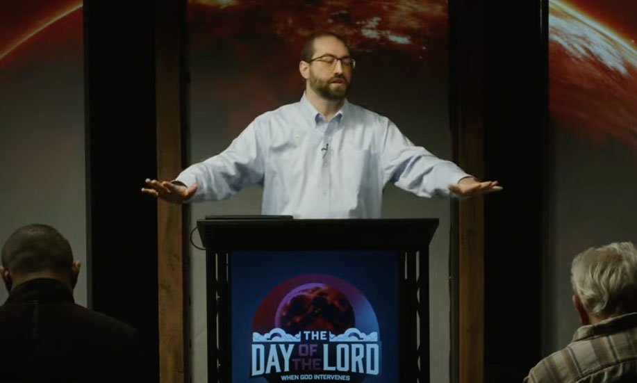 A man with glasses and a beard stands behind a podium, speaking or giving a presentation to an audience in a church or conference room. There are people seated and a screen behind him displaying an image of Earth with the text "The Day of the Lord."