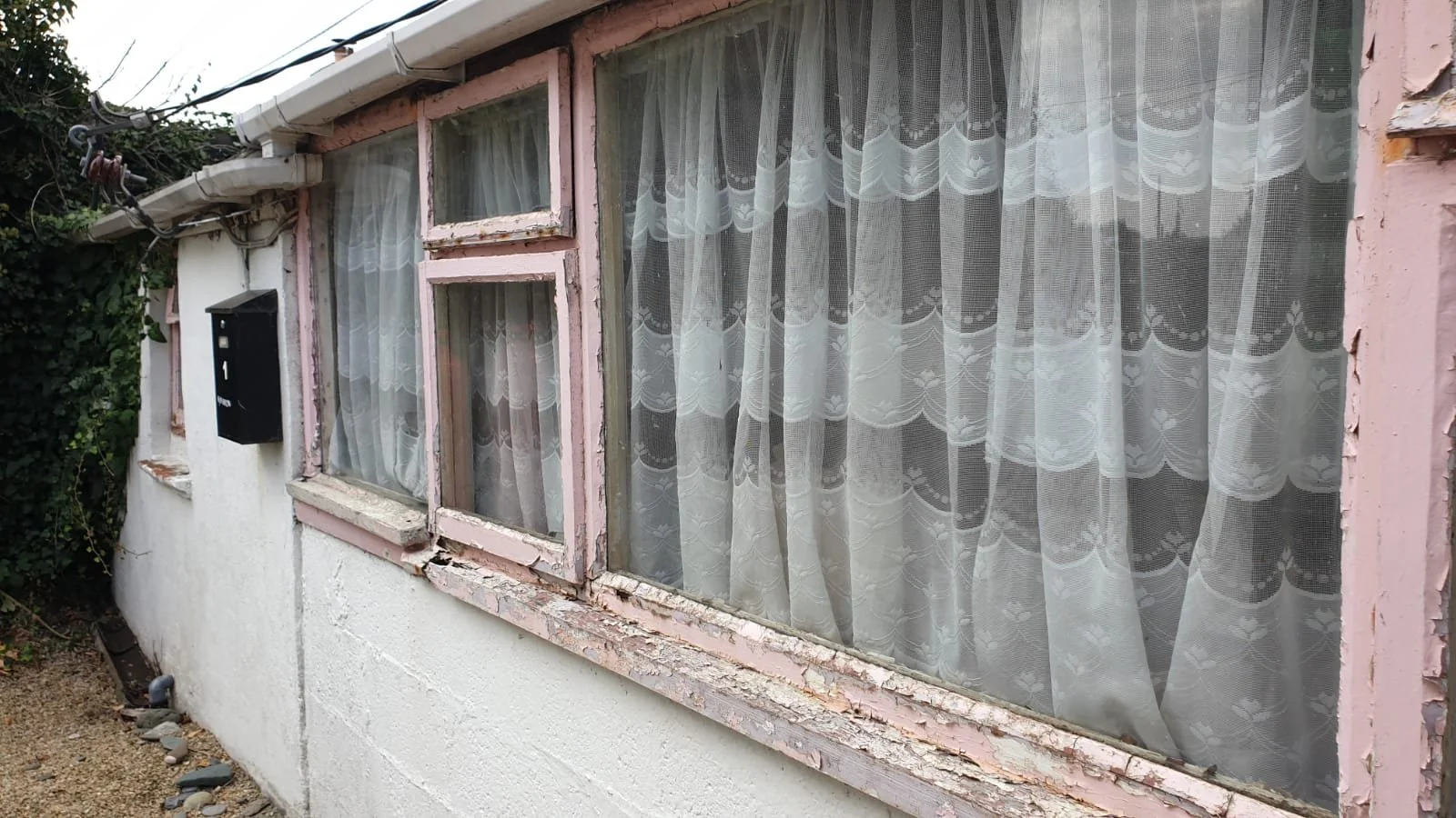 pink timber rotted window frames and white render and overgrown bushes