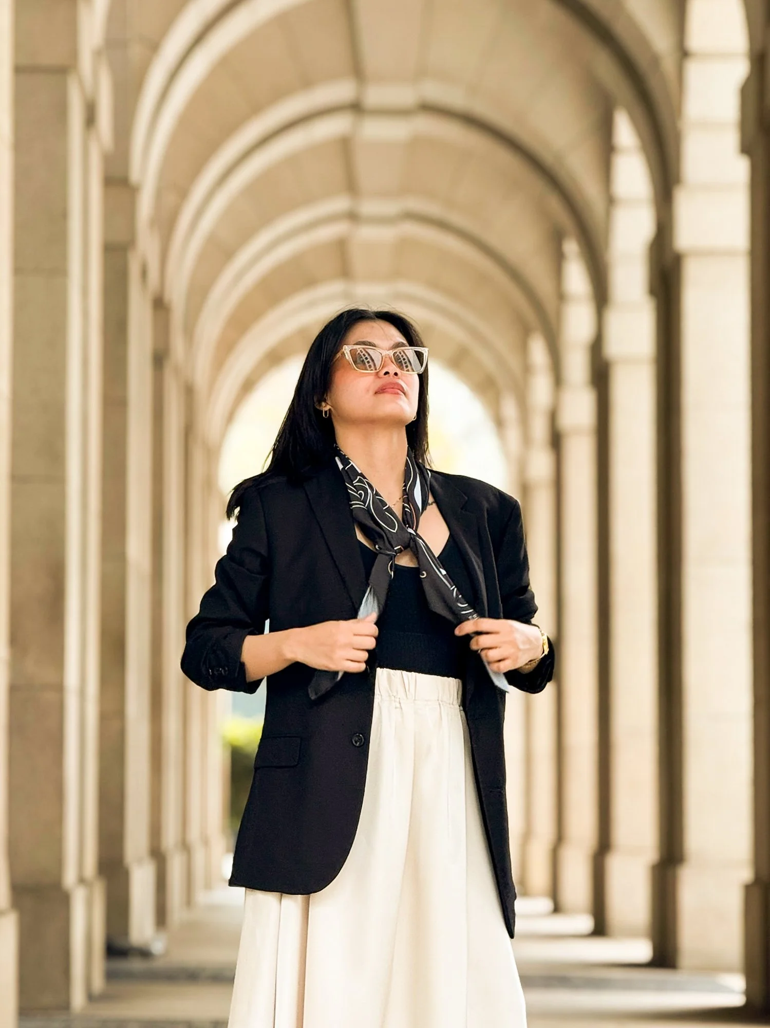 A woman wearing sunglasses, a black blazer, a black and white scarf, and white pants, standing in an arched outdoor corridor.