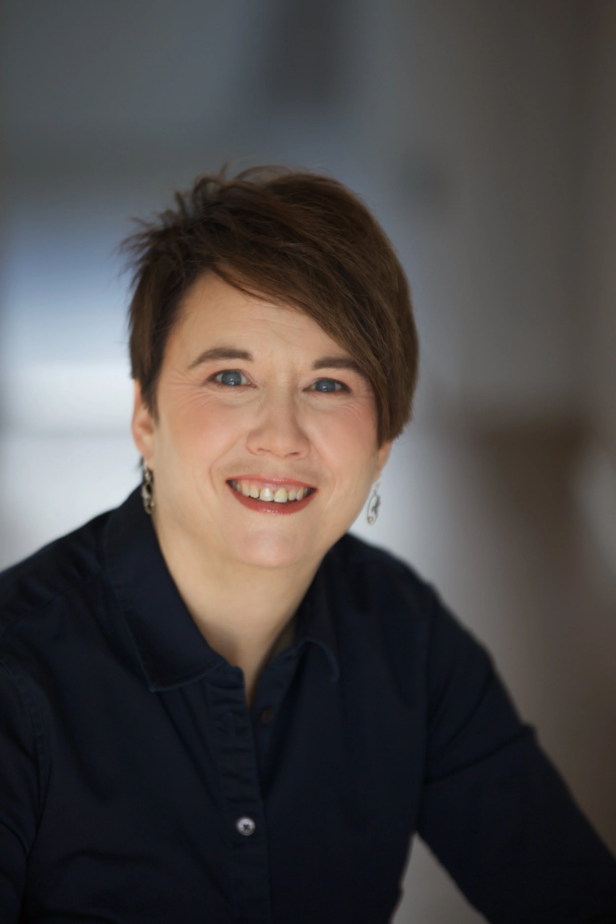 Portrait of a woman with short brown hair, wearing a black and white patterned blouse, smiling against a plain light background. This is the Managing Director of WorldUpstart.