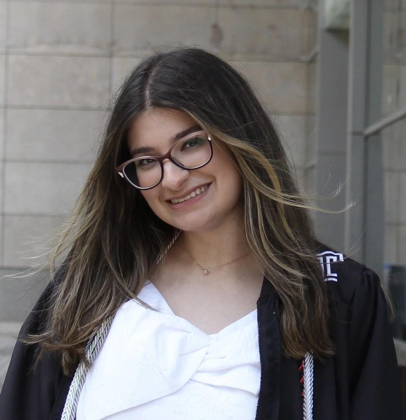 A young woman with long brown hair, wearing glasses, a white top, and a black graduation gown, smiling outdoors. This is WorldUpstart's Communication and Program Lead, Brianna Amato.