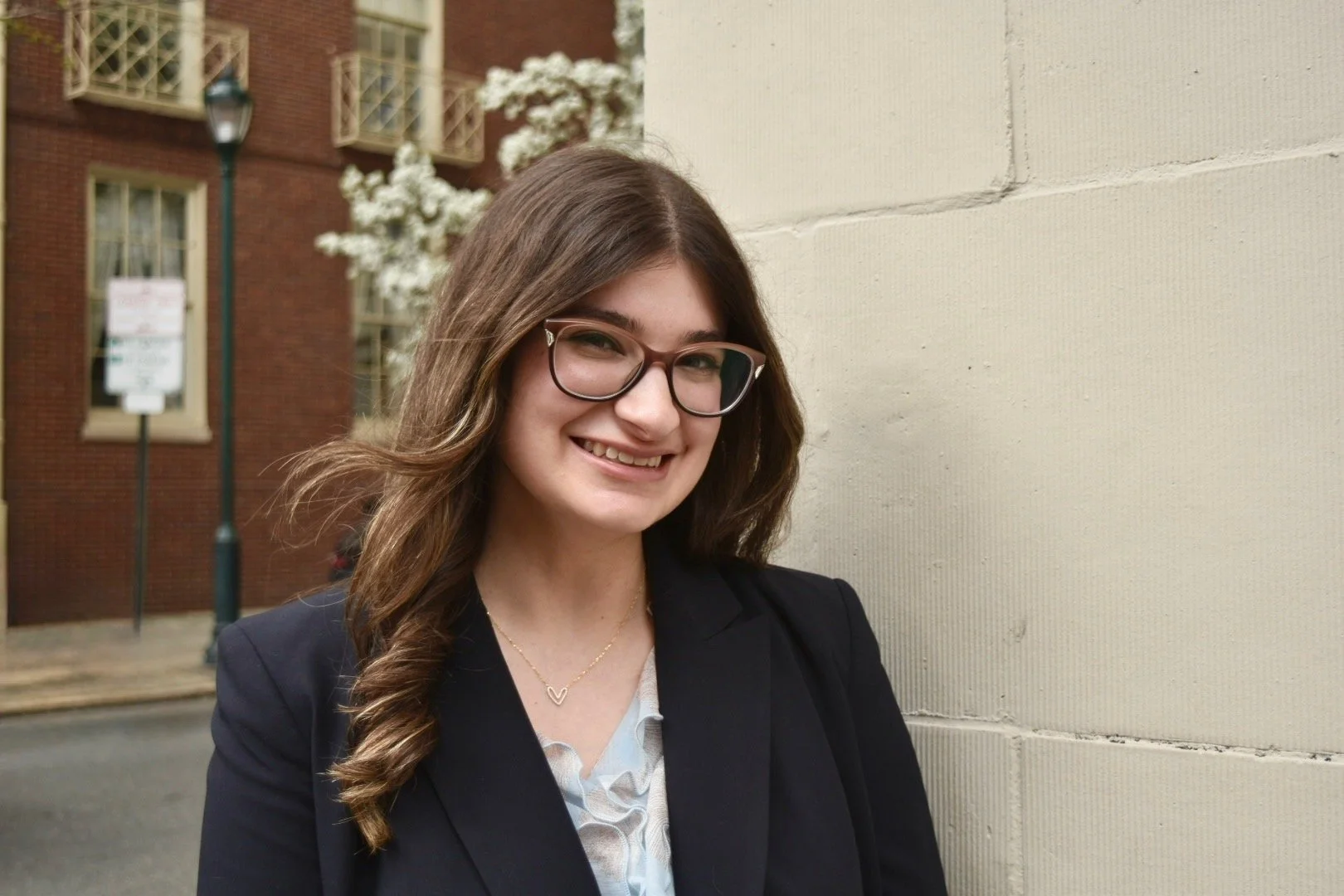A young woman with long brown hair, wearing glasses, a white top, and a black graduation gown, smiling outdoors. This is WorldUpstart's Communication and Program Lead, Brianna Amato.