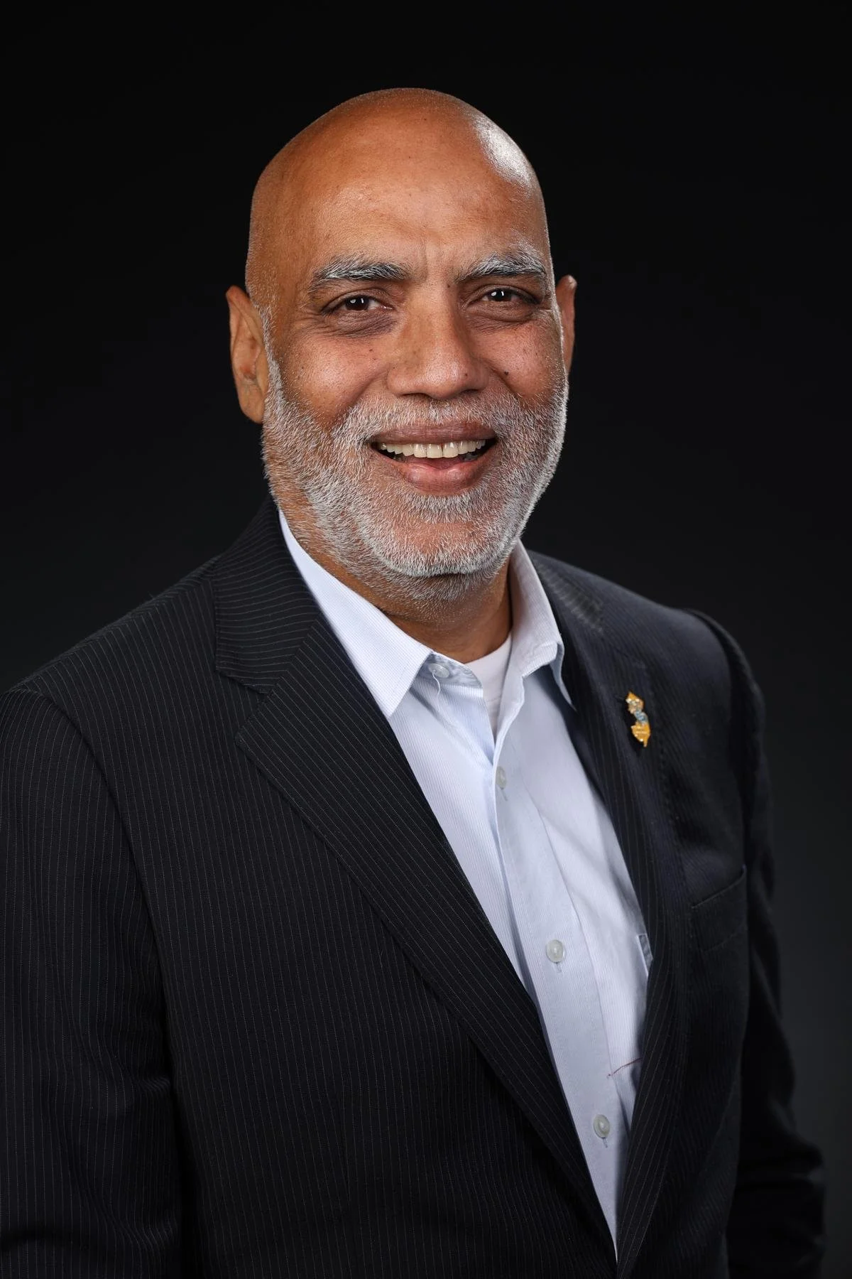 A man with a bald head and gray beard, wearing a dark pinstripe suit and a light blue dress shirt, standing with arms crossed against a plain gray background. This is WorldUpstart's mentor, Sunil Joshi.
