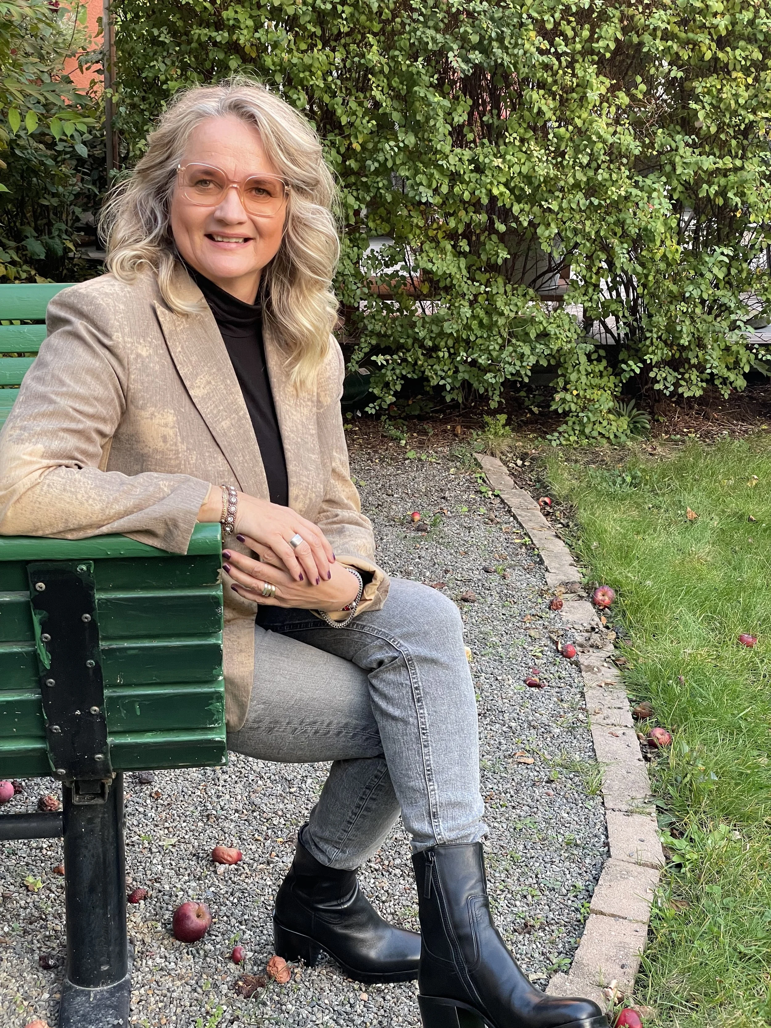 En kvinna med blont, lockigt hår, glasögon, gul skjorta, svarta byxor och smycken sitter i en ljus fåtölj och pratar eller förklarar.