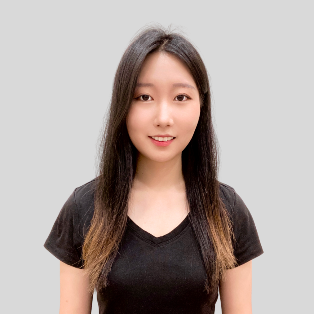 A young woman with long dark hair smiling, wearing a black V-neck T-shirt, against a plain gray background.
