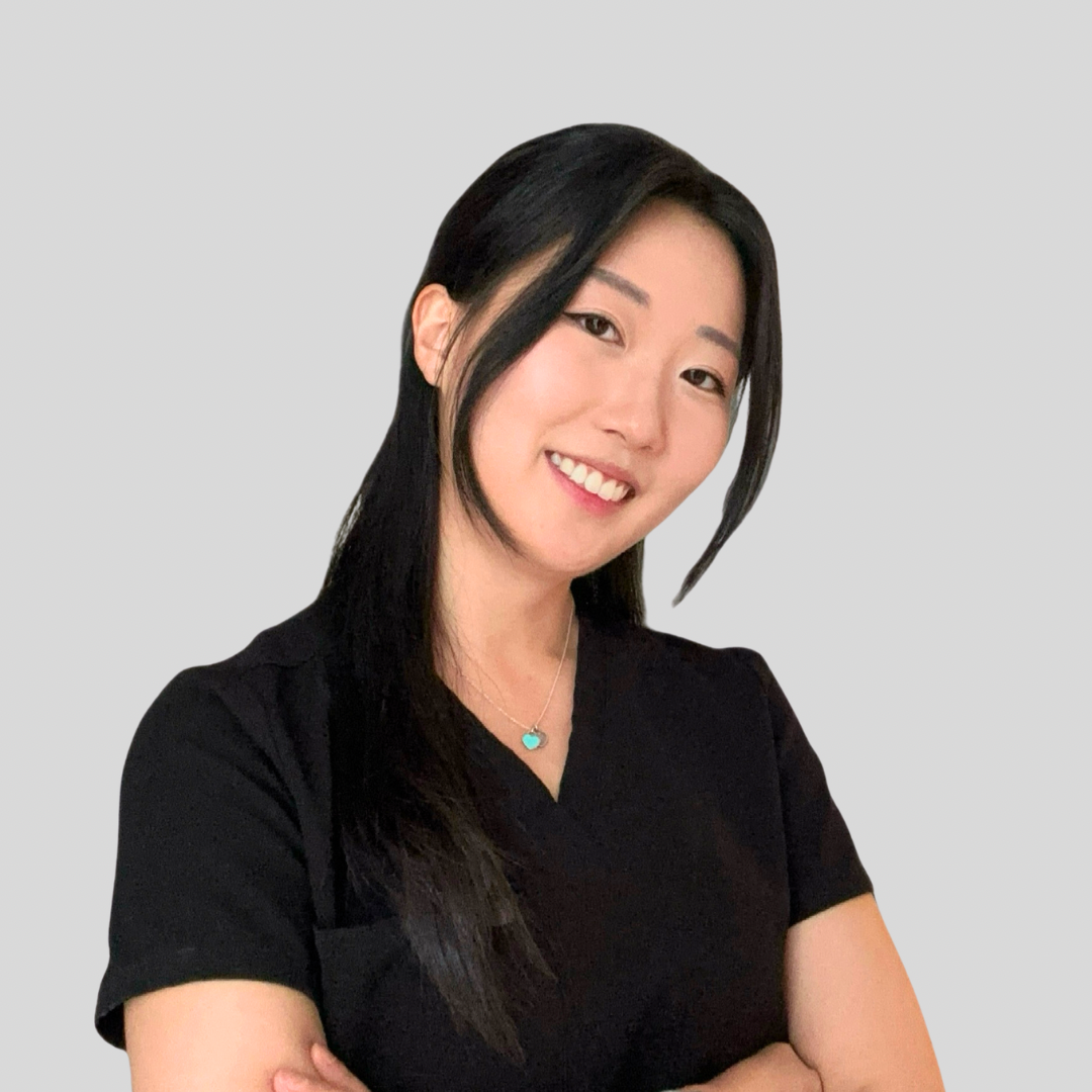 A young woman with long dark hair smiling, wearing a black V-neck T-shirt, against a plain gray background.