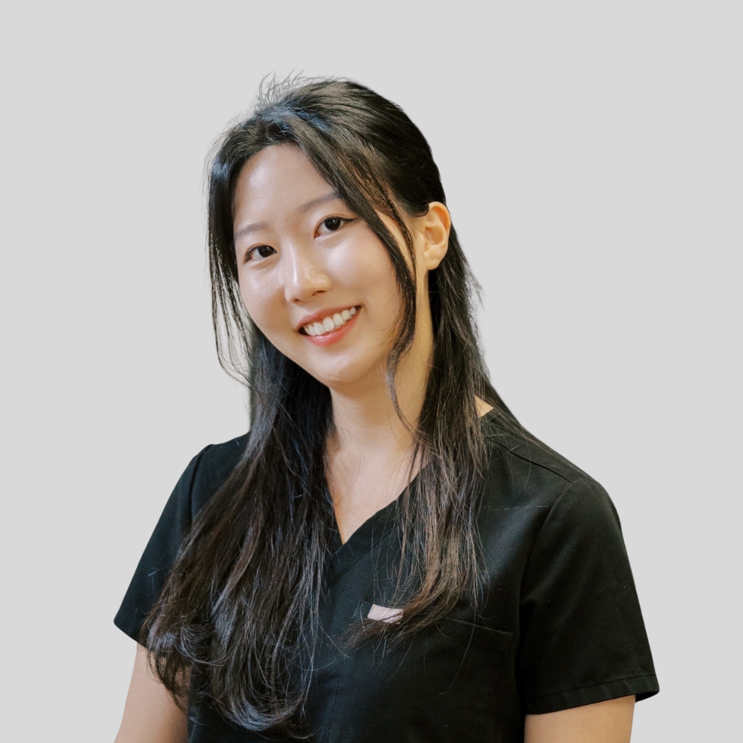 A young woman with long dark hair smiling, wearing a black V-neck T-shirt, against a plain gray background.