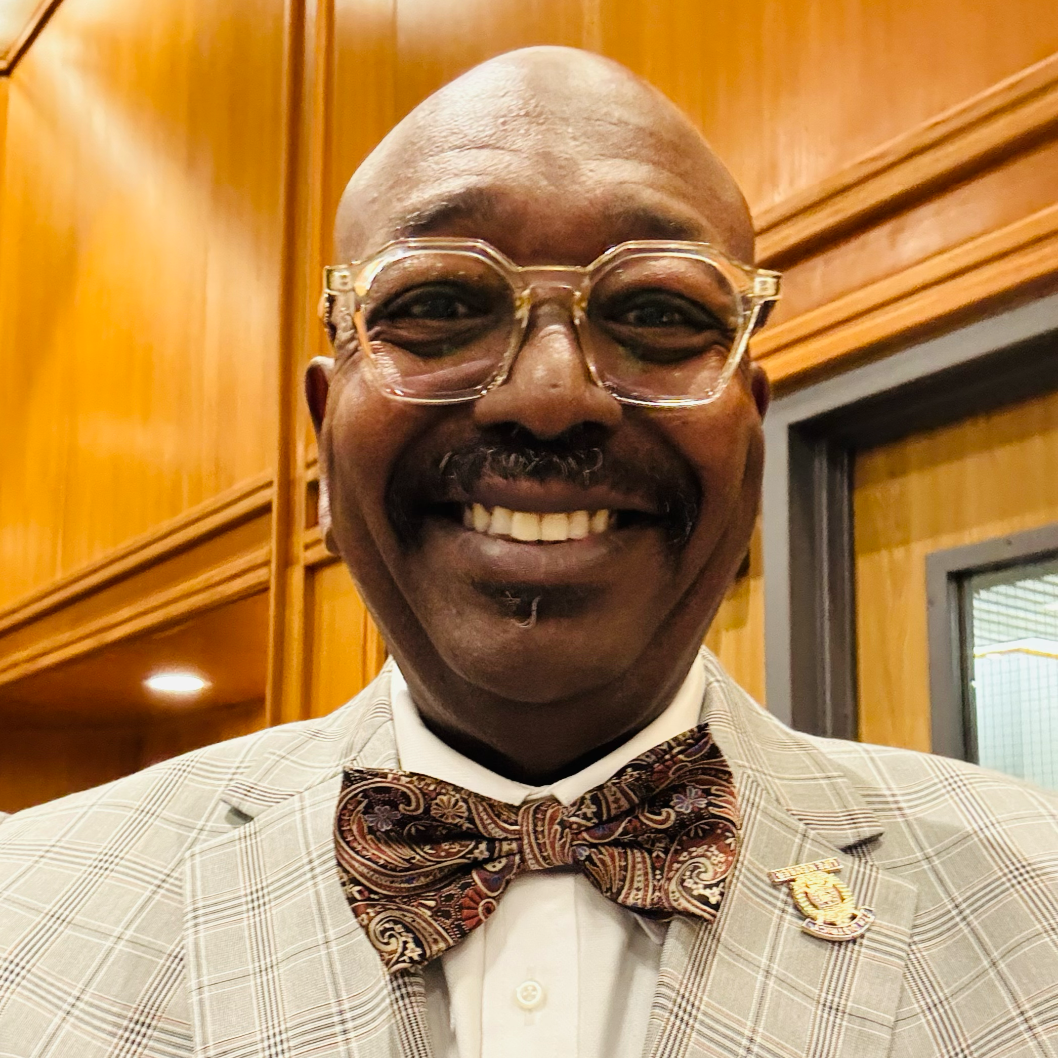 Man in a suit with glasses, wearing a patterned tie, smiling in a circular frame.