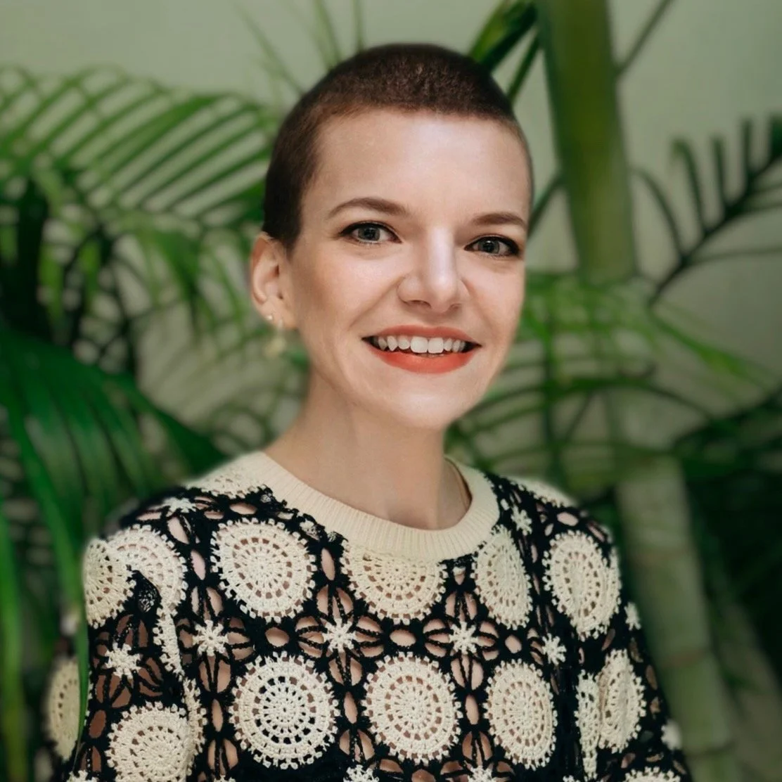 A young woman with short hair and a warm smile, wearing a black and white crochet patterned sweater, standing in front of green leafy plants.