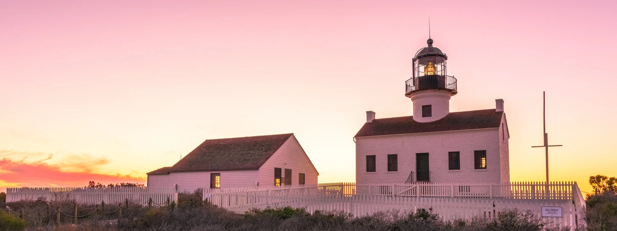 Panoramic view of the white Old Point Loma Lighthouse and keeper's quarters in San Diego, California, sitting behind a white picket fence under a vibrant pink and purple sunset sky with the lantern room illuminated.