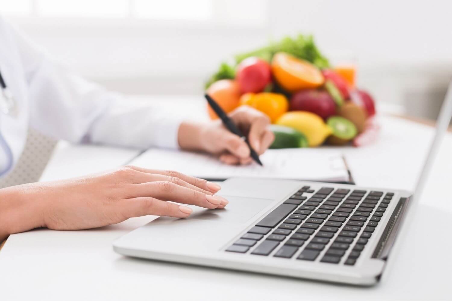 Persona tecleando en una laptop con un cuaderno y un bolígrafo cerca, y un arreglo de frutas en el fondo.