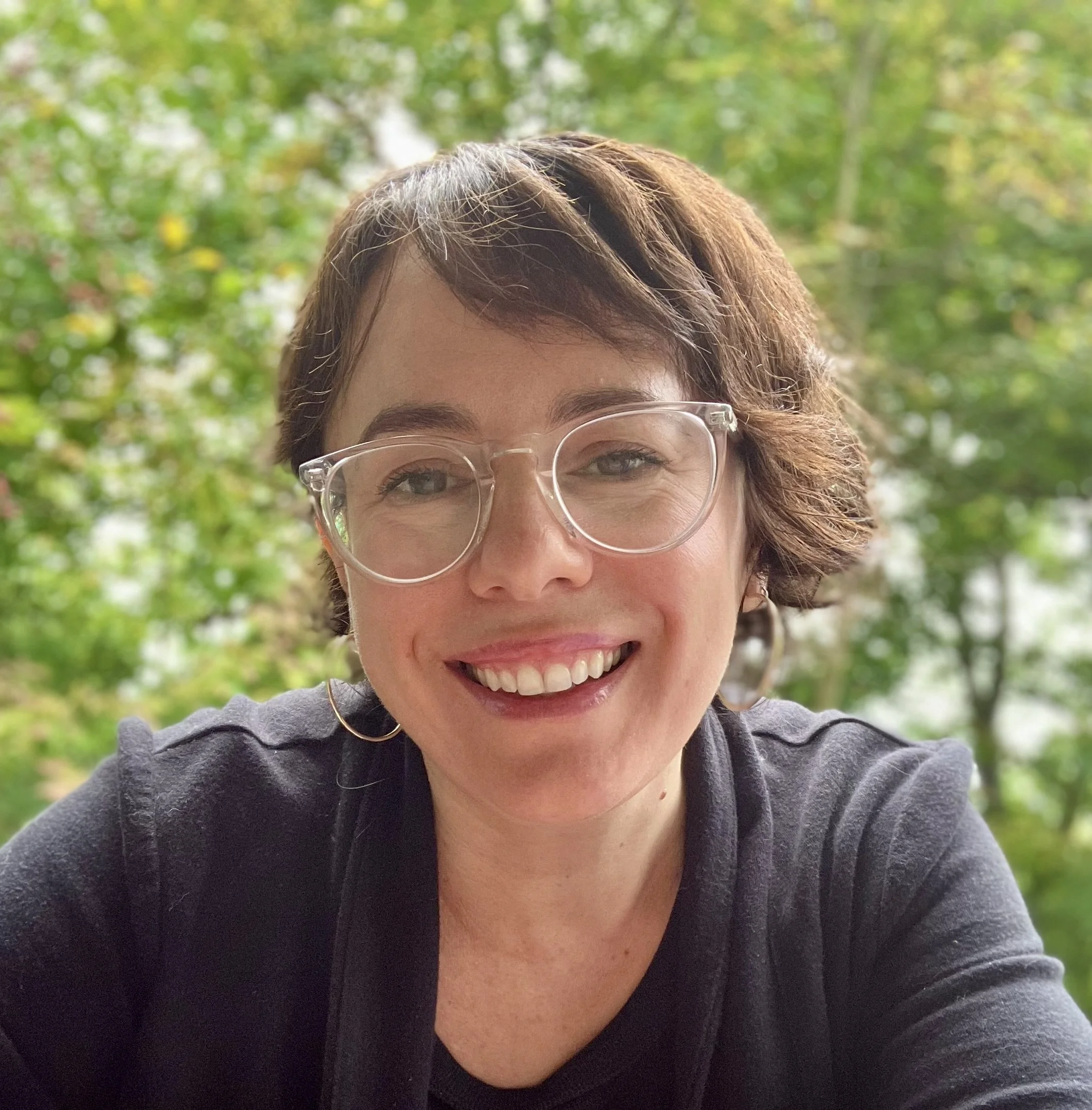 A smiling woman with short brown hair and glasses outdoors in a wooded area.