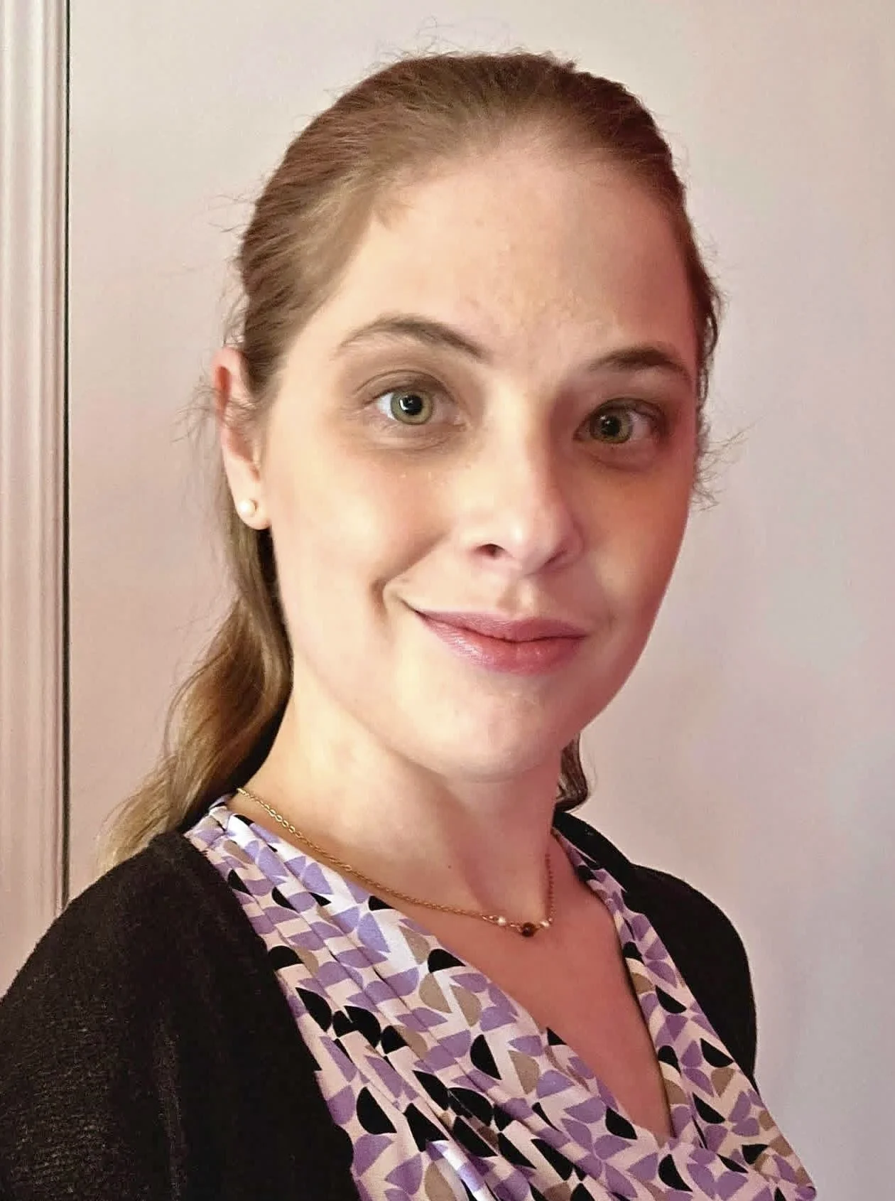 A young woman with light brown hair tied back, wearing a patterned blouse, a gold chain necklace, and small pearl earrings, standing against a light-colored wall.