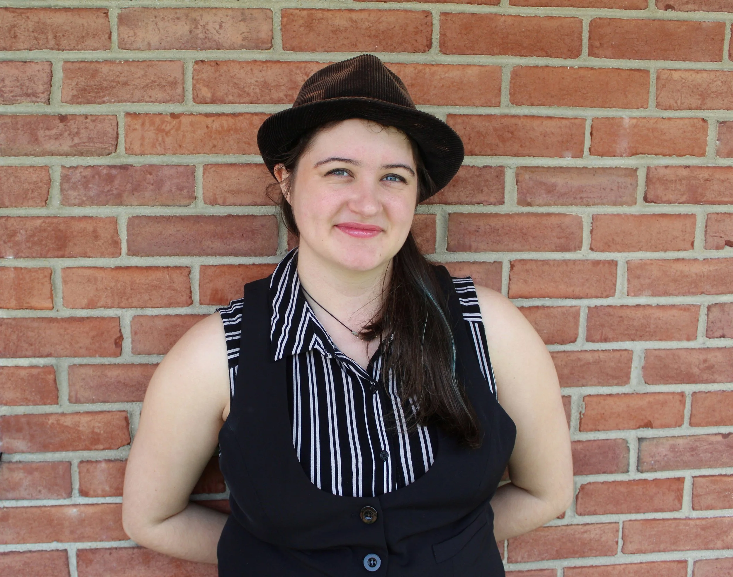 Young woman with long dark hair, wearing a black sleeveless vest over a black and white striped shirt and a dark brown hat, standing against a brick wall.