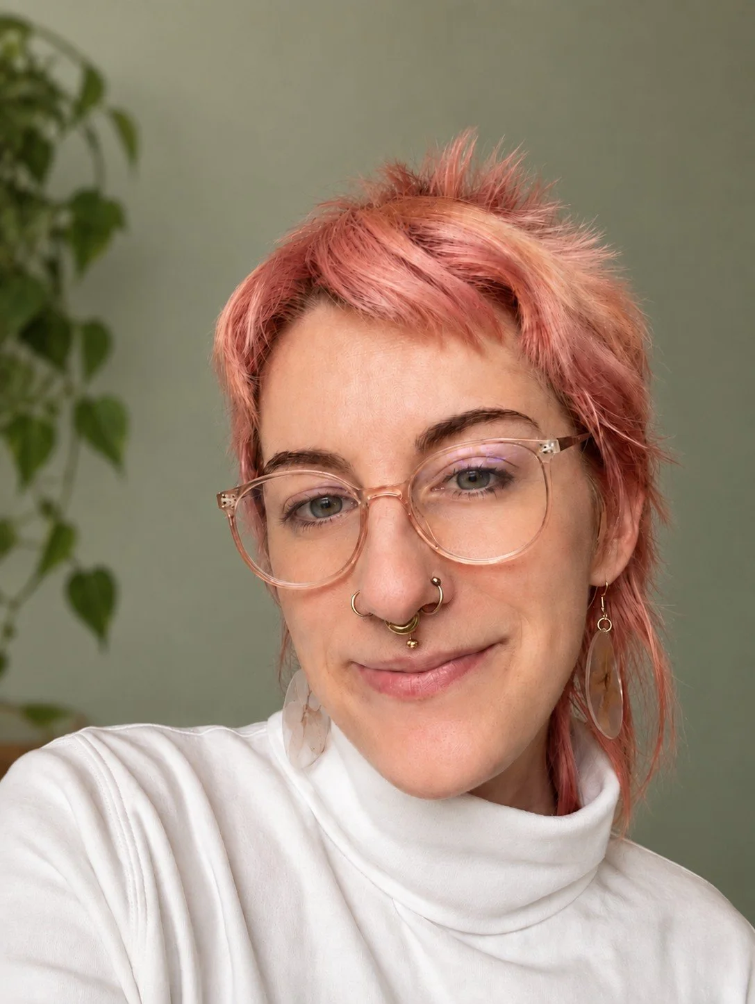 Libby Farren, a therapist with short pink hair, clear-framed glasses, and a white turtleneck, smiling in front of a sage green wall with a plant.