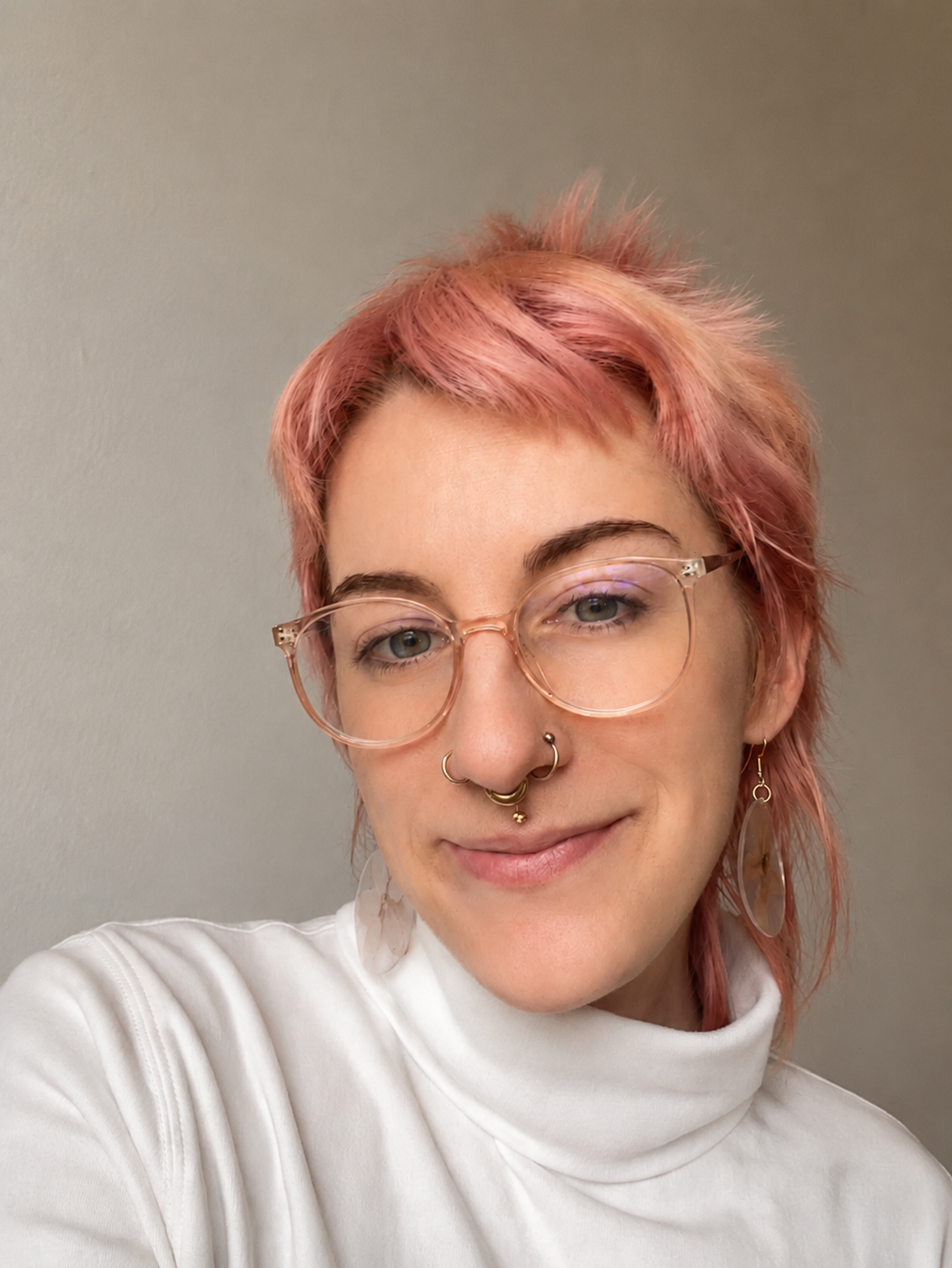 A woman with short pink hair, glasses, and multiple piercings smiling in front of a plain wall.