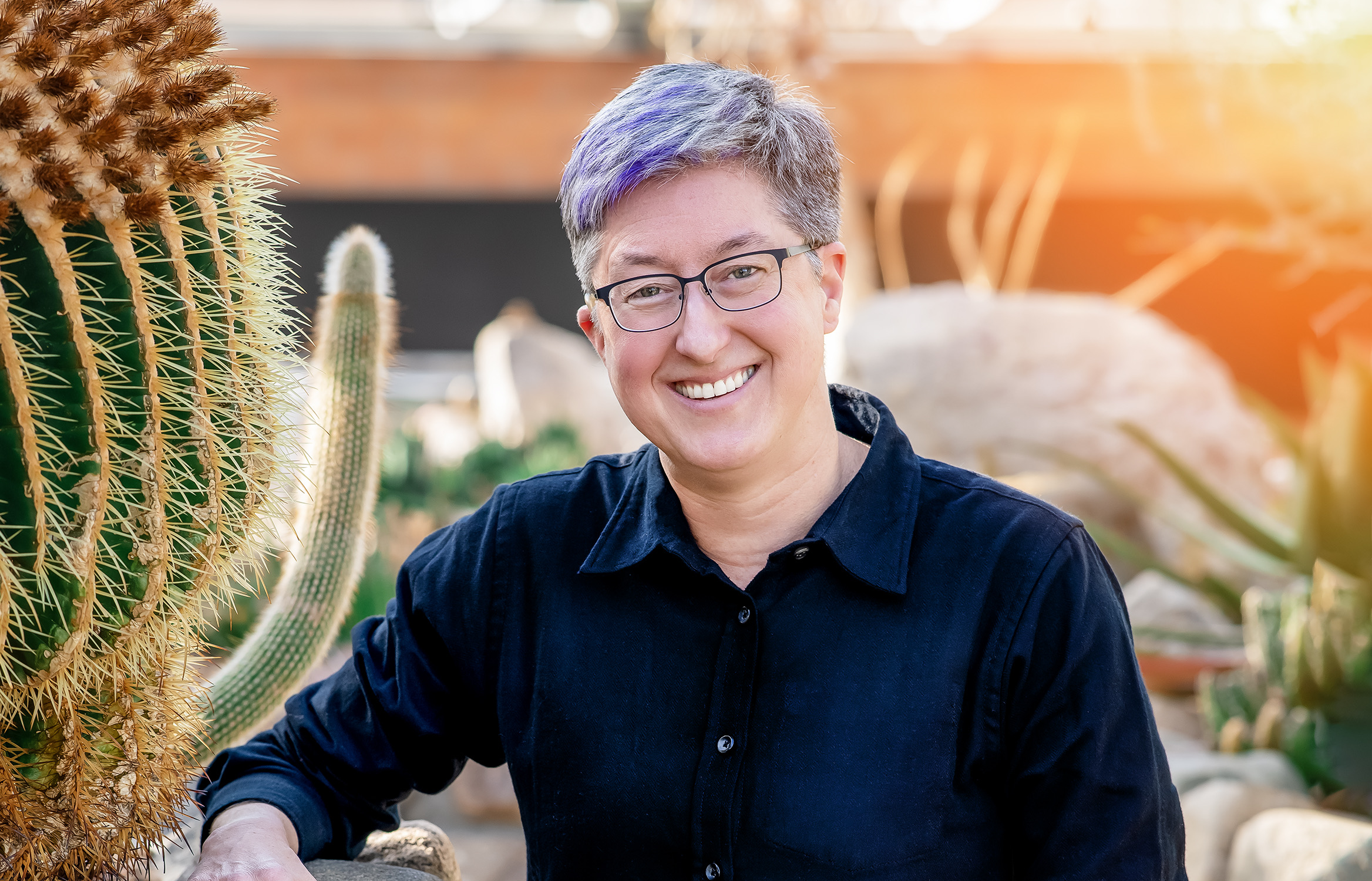A person smiling and wearing glasses standing in a cactus garden during the daytime.