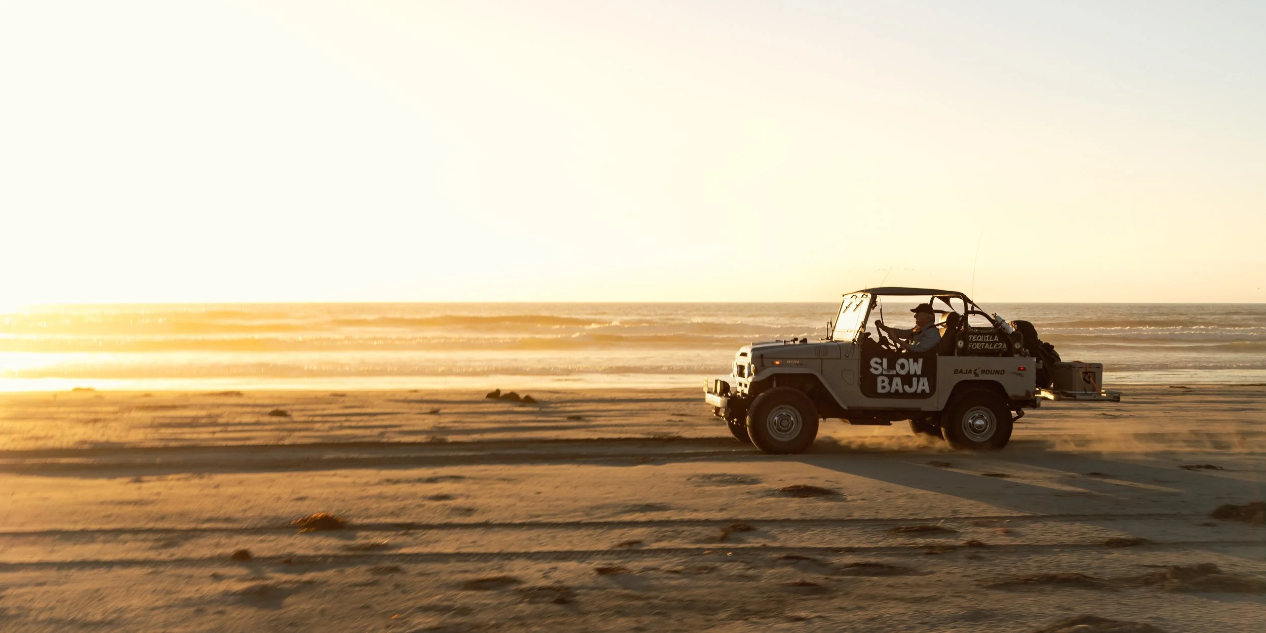 Slow Baja expedition vehicle driving along a beach at sunset, with Slow Baja lettering on its side