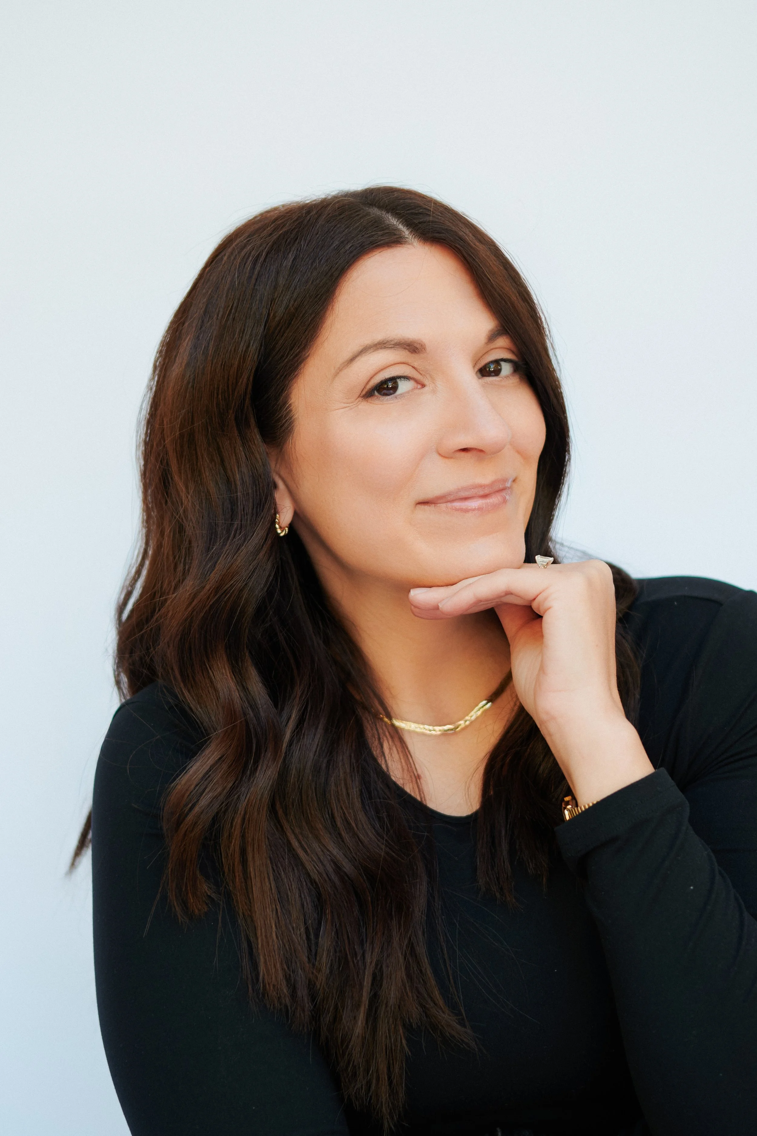 Close-up portrait of a smiling woman with long brown hair, gold jewelry, and her hand resting under her chin