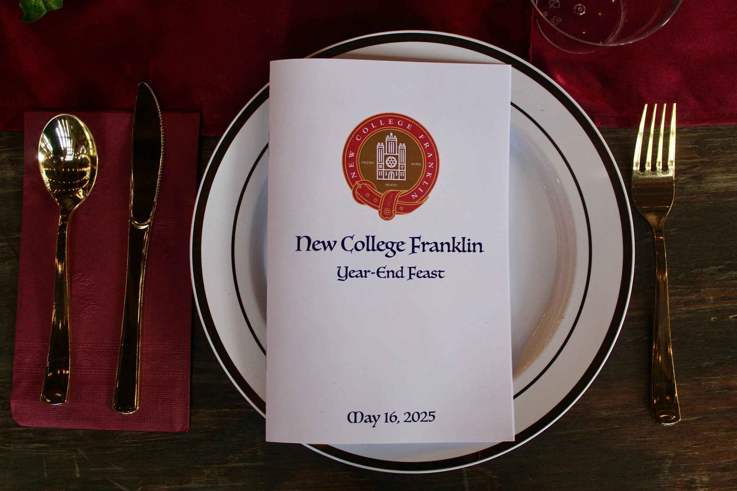 Table setting with gold utensils, a white plate with black rim, a maroon napkin, a glass of water, and a program card for New College Franklin Year-End Feast dated May 16, 2025, featuring a red and gold college emblem.