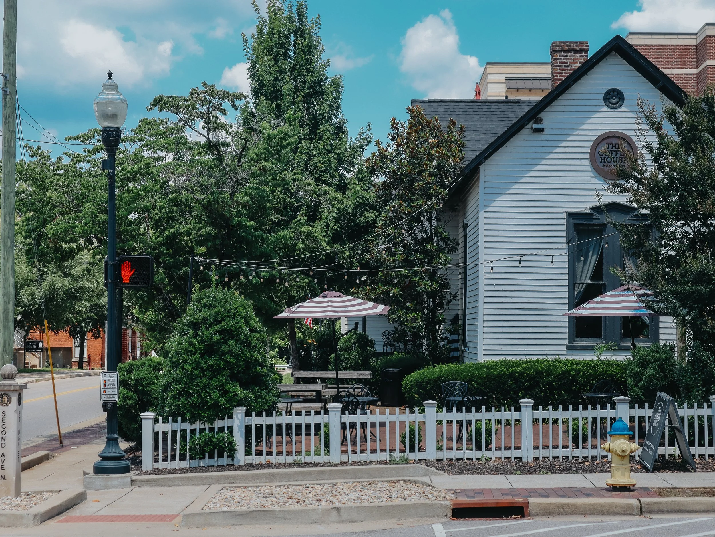 A small outdoor seating area with tables and umbrellas in front of a white wooden building labeled 'The Coffee House'. There are trees, greenery, and a street with a traffic light showing a red hand signal.