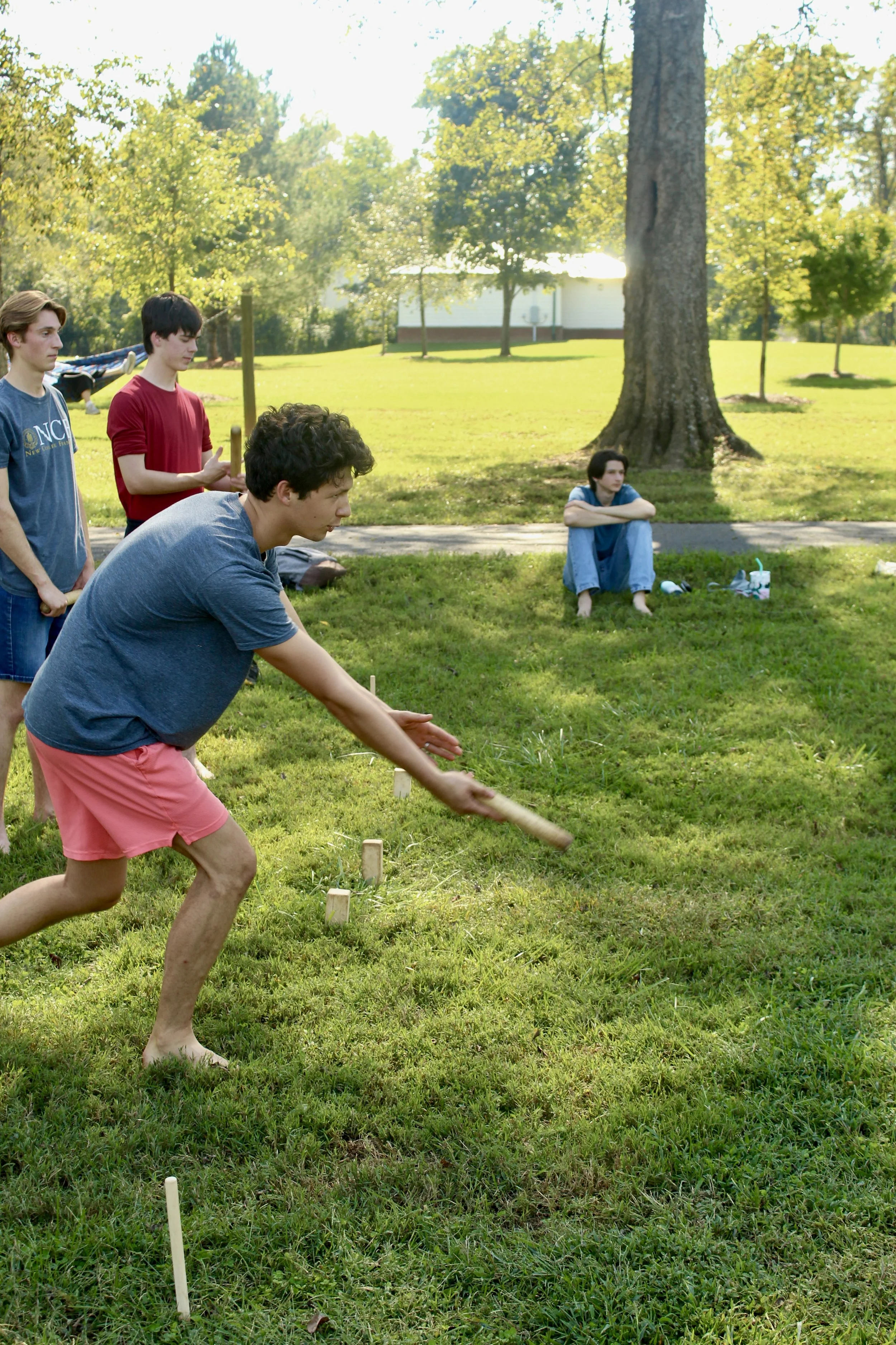 Four young men playing cornhole outdoors on a grassy lawn near trees, with one sitting on the grass and the other three standing or leaning, during a sunny day.