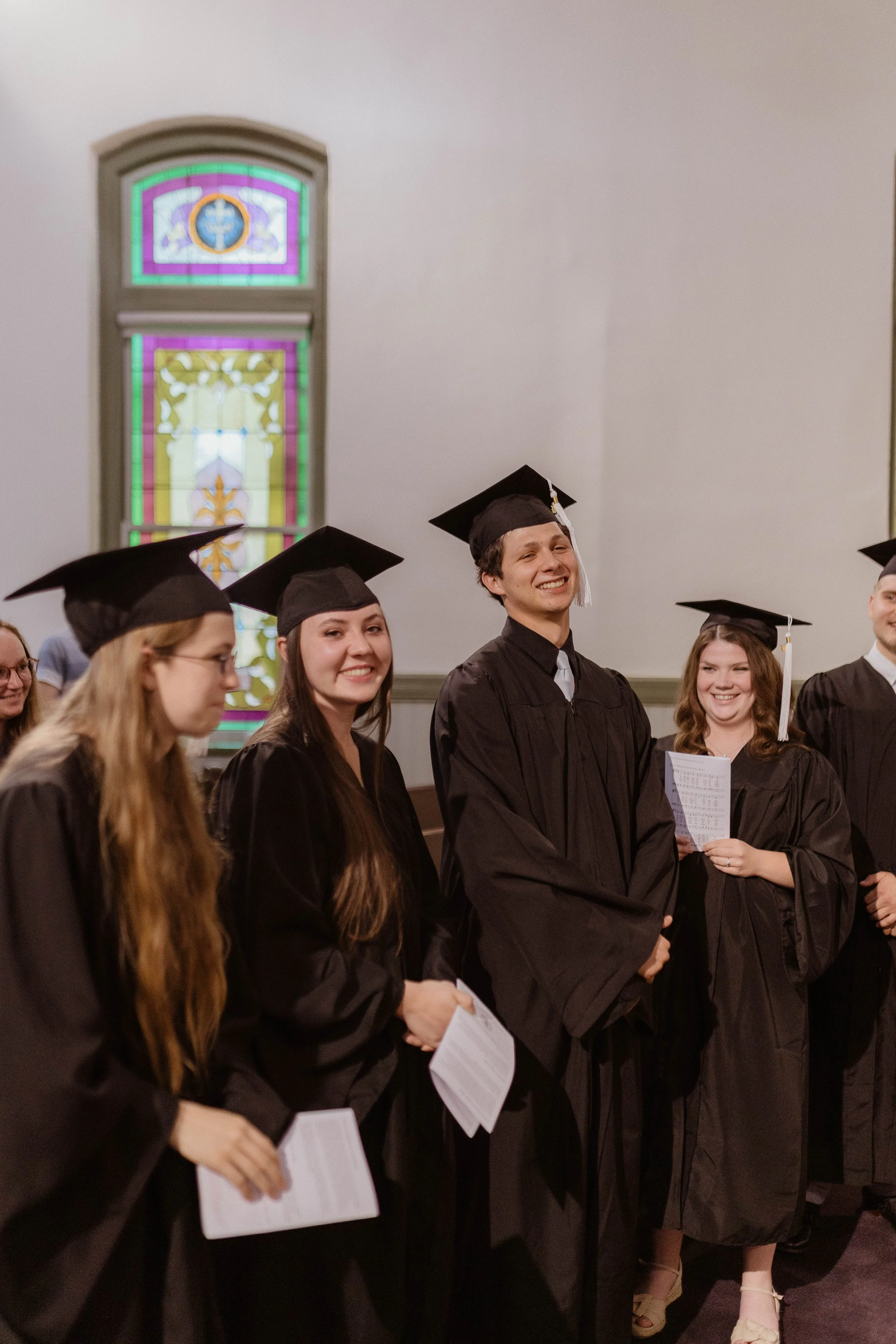 Group of students in black graduation gowns and caps during a graduation ceremony inside a church with stained glass windows.