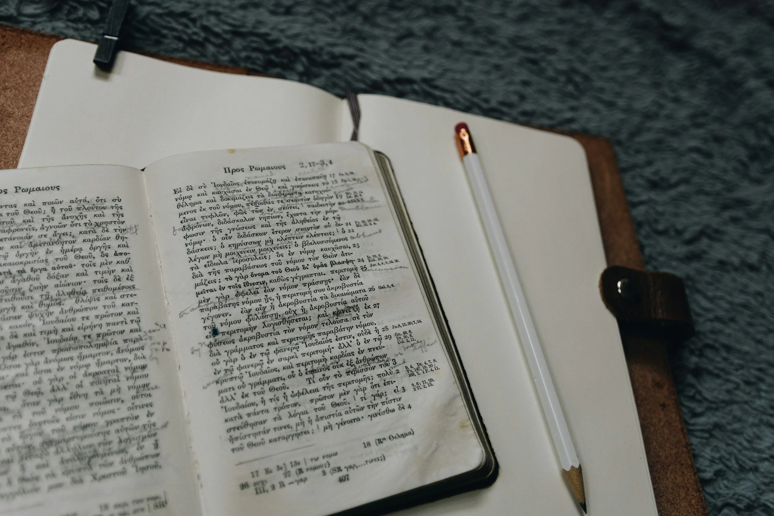 Open book with Greek text on a desk, a pencil nearby, and water in the background.