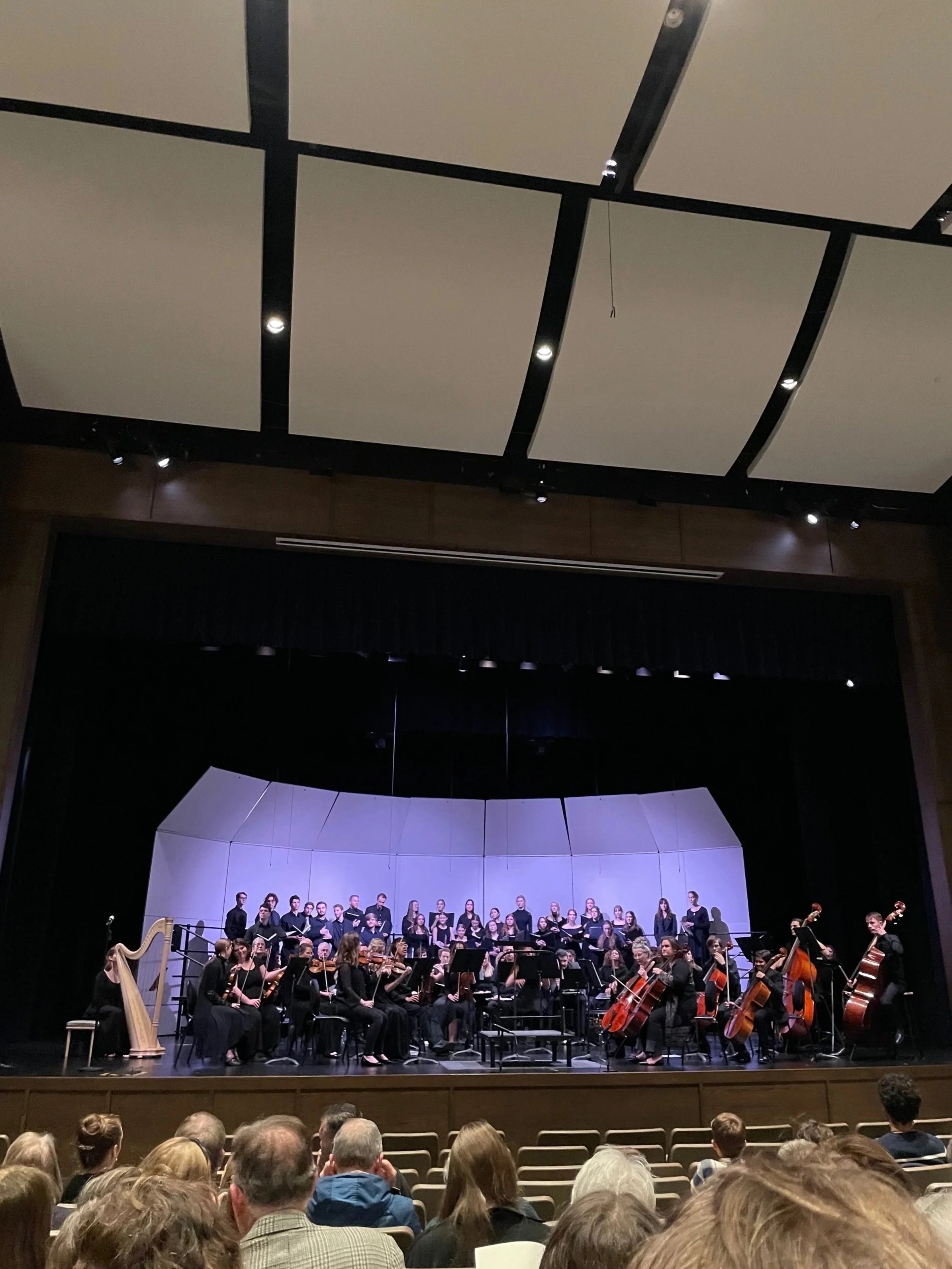 Orchestra performing on stage with a choir in the background, audience seated in front.