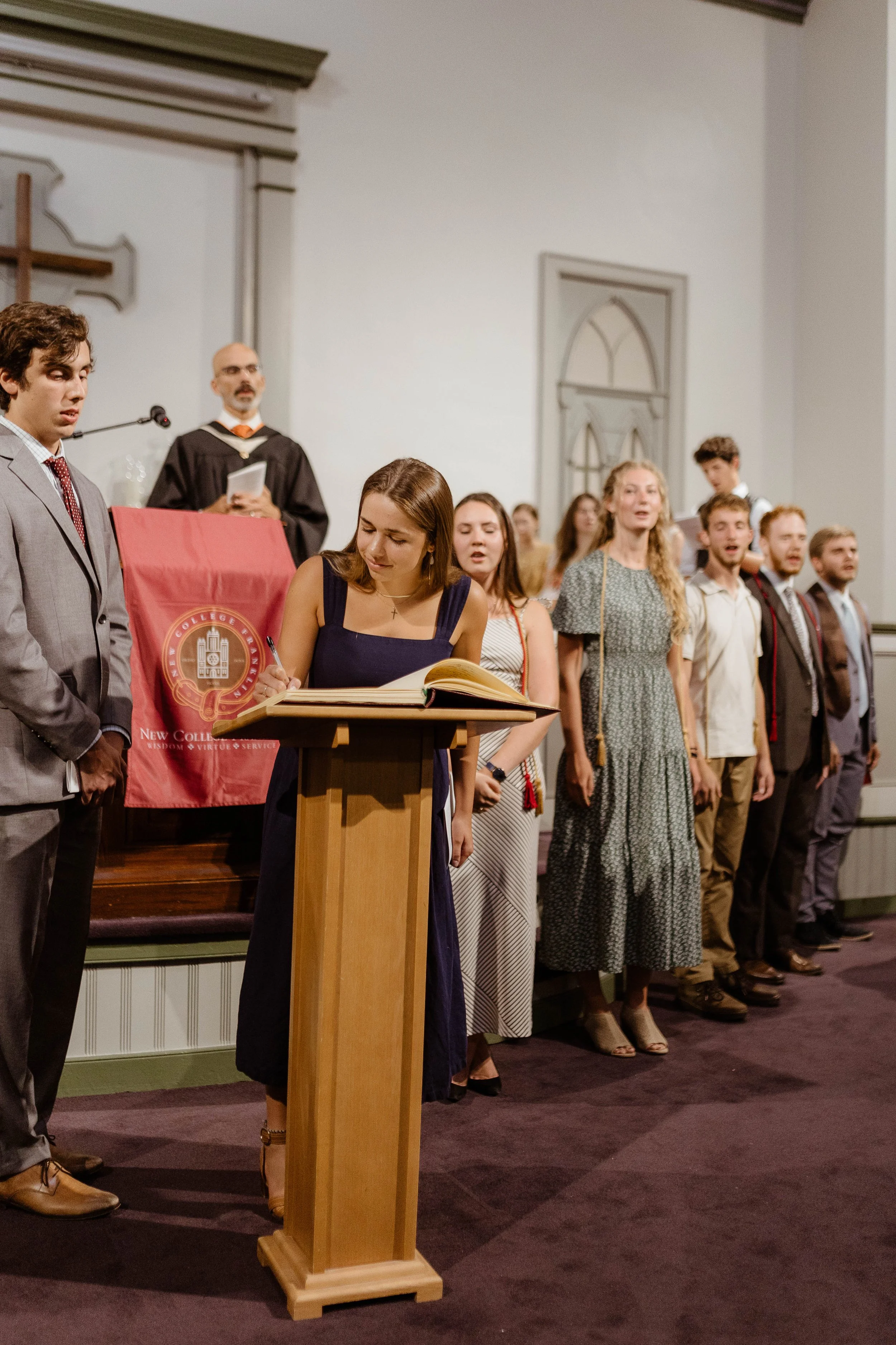 People standing in a line at a ceremony, with a woman signing at a podium and a man in a graduation gown in the background.