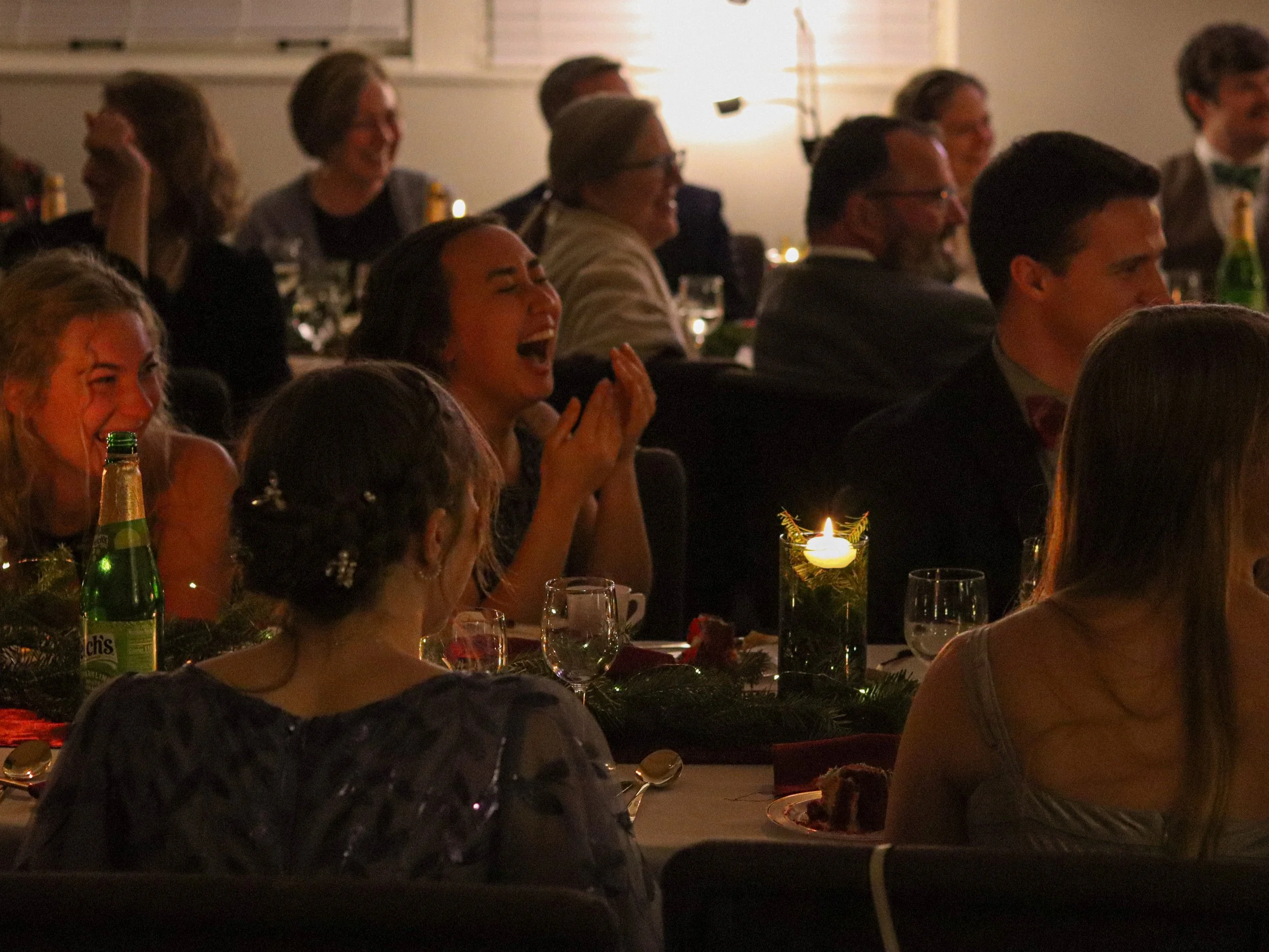 People laughing and enjoying a festive gathering around a decorated table with candles and drinks in a dimly lit room.