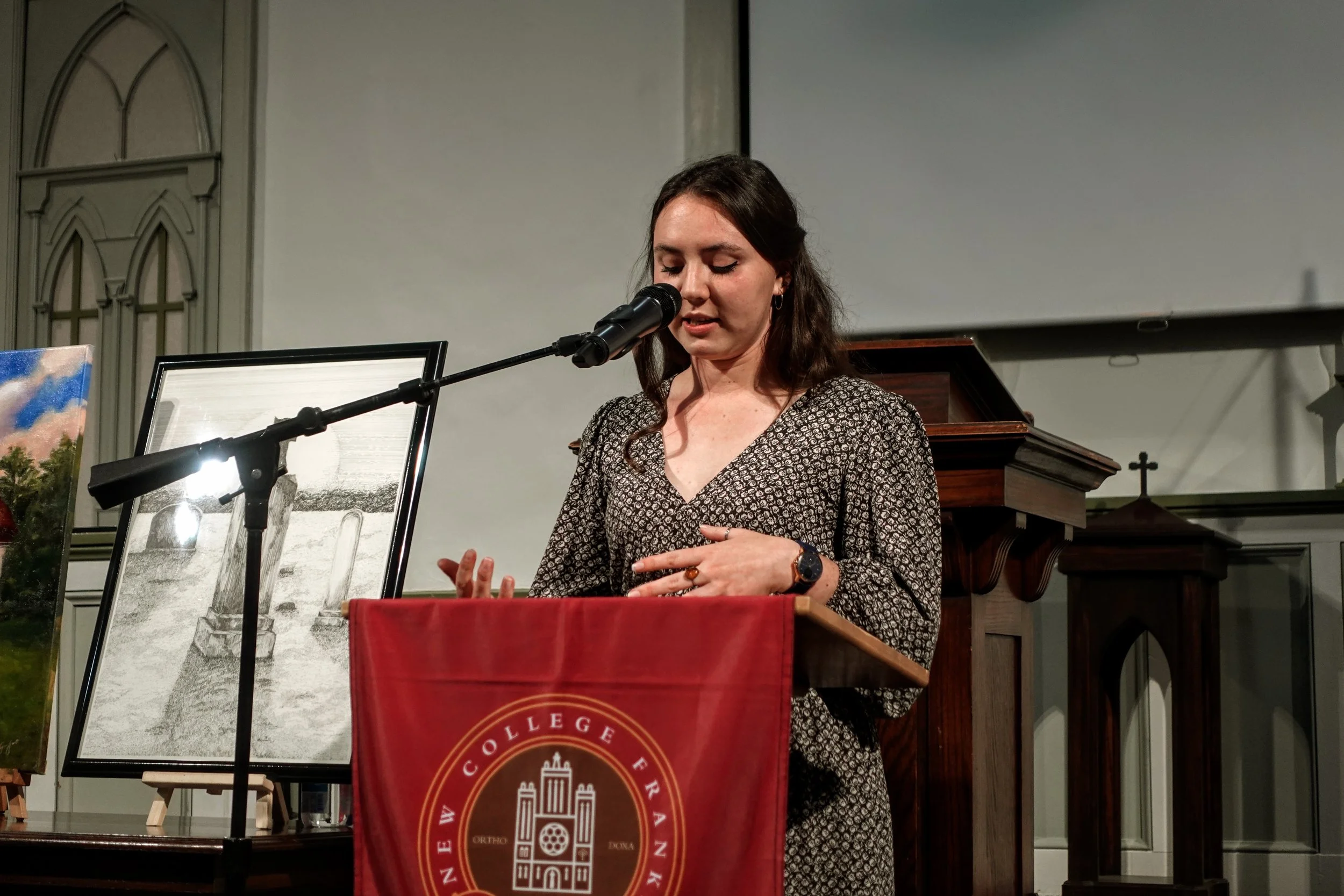 A woman with long brown hair, wearing a patterned black and white dress, is standing at a podium with a red cloth featuring a gold emblem. She appears to be speaking or reading into a microphone inside a building, possibly a church or hall, with framed pictures and artwork on display nearby.
