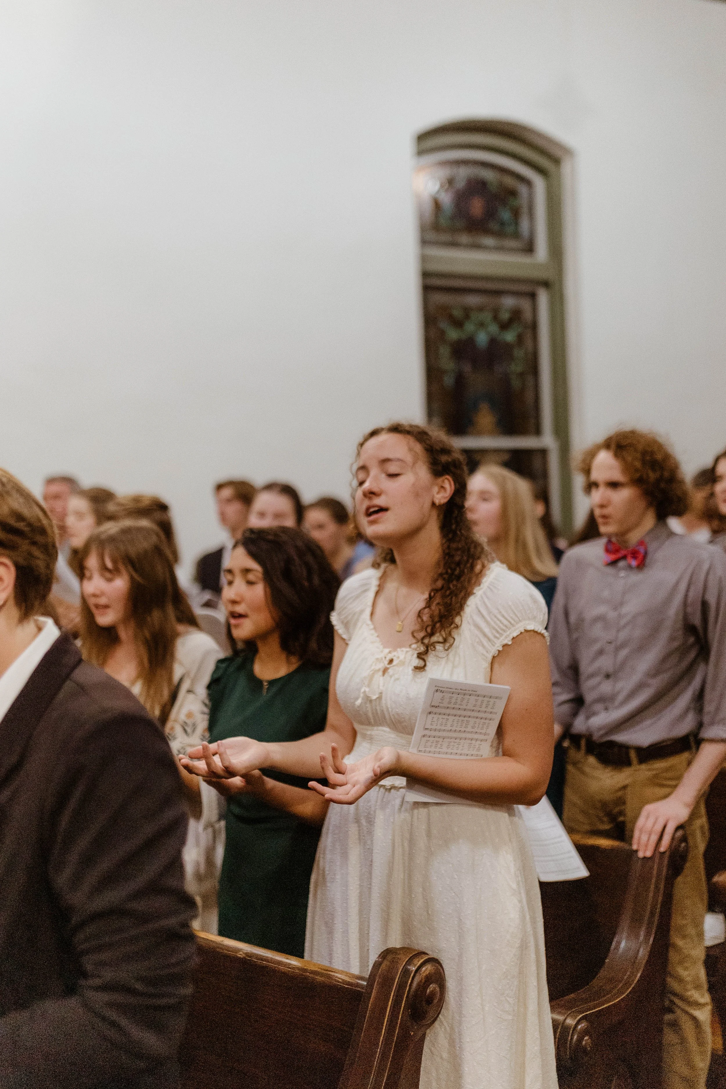 A woman in a white dress with wavy hair holding a program book, standing in a church during a religious ceremony with people behind her.