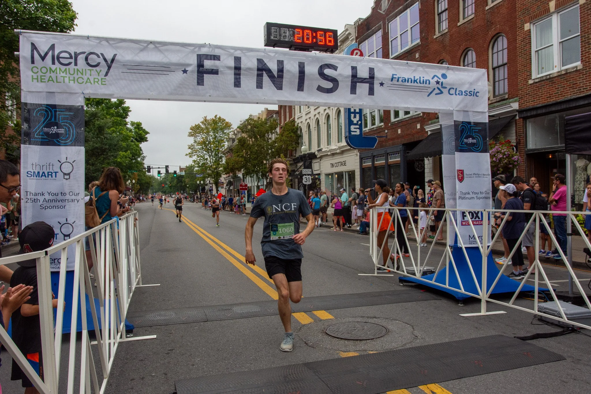 A runner crossing the finish line at the Franklin Classic race, with a crowd of spectators and buildings in the background on a cloudy day.