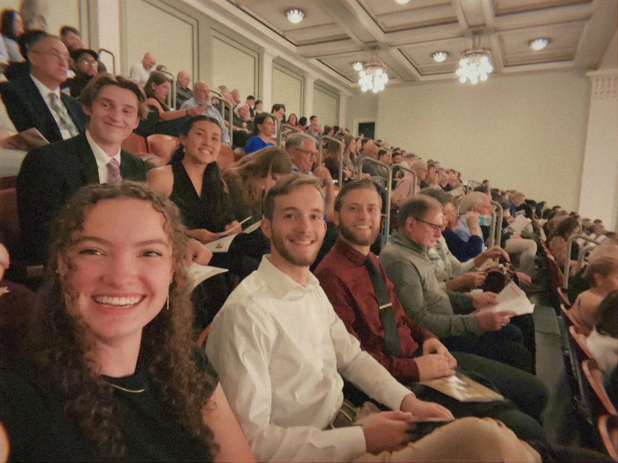 Photo of a large group of diverse people seated in an auditorium, attending an event or conference, with some smiling at the camera.
