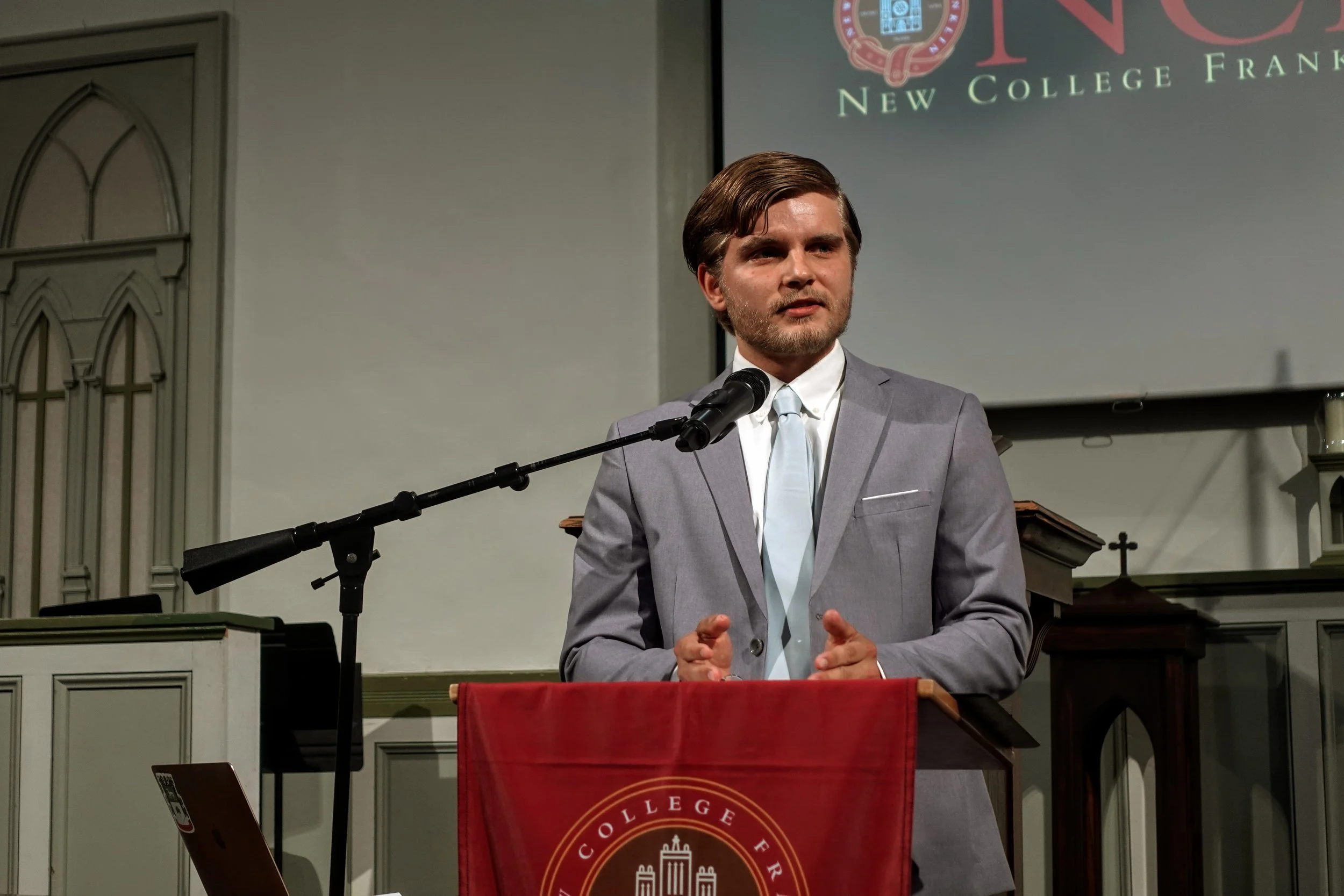 A man in a gray suit and light blue tie speaking at a liberal arts school.