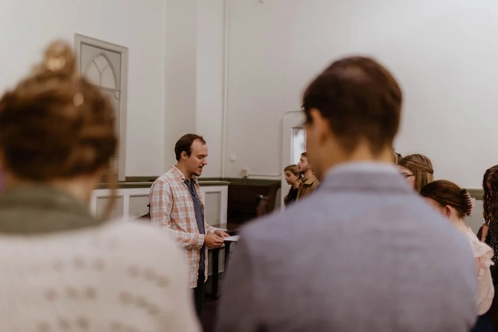 A man standing at the front of a room, reading from a paper, with a group of people standing in rows, listening.