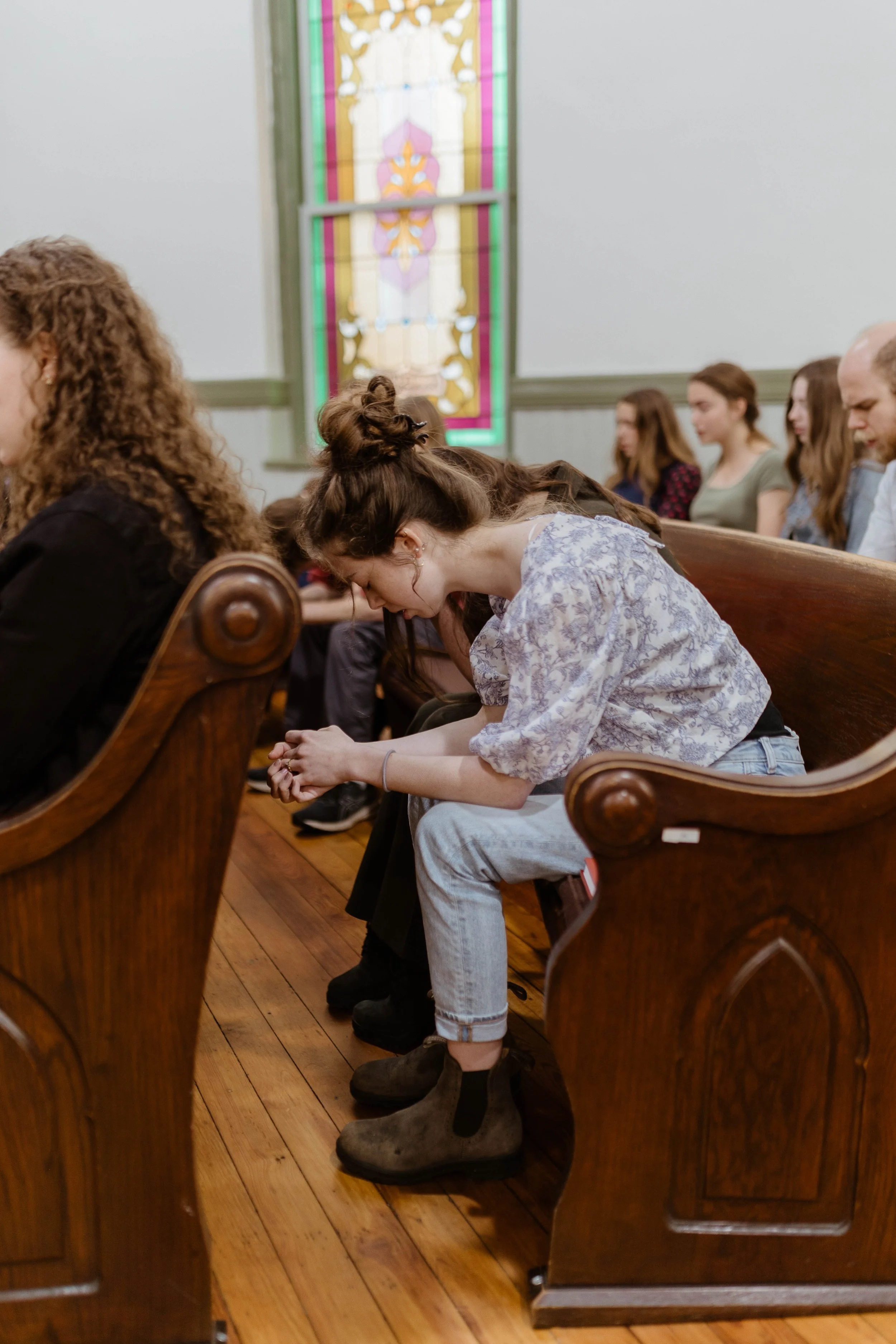 People sitting on wooden pews in a church, with a stained glass window in the background.