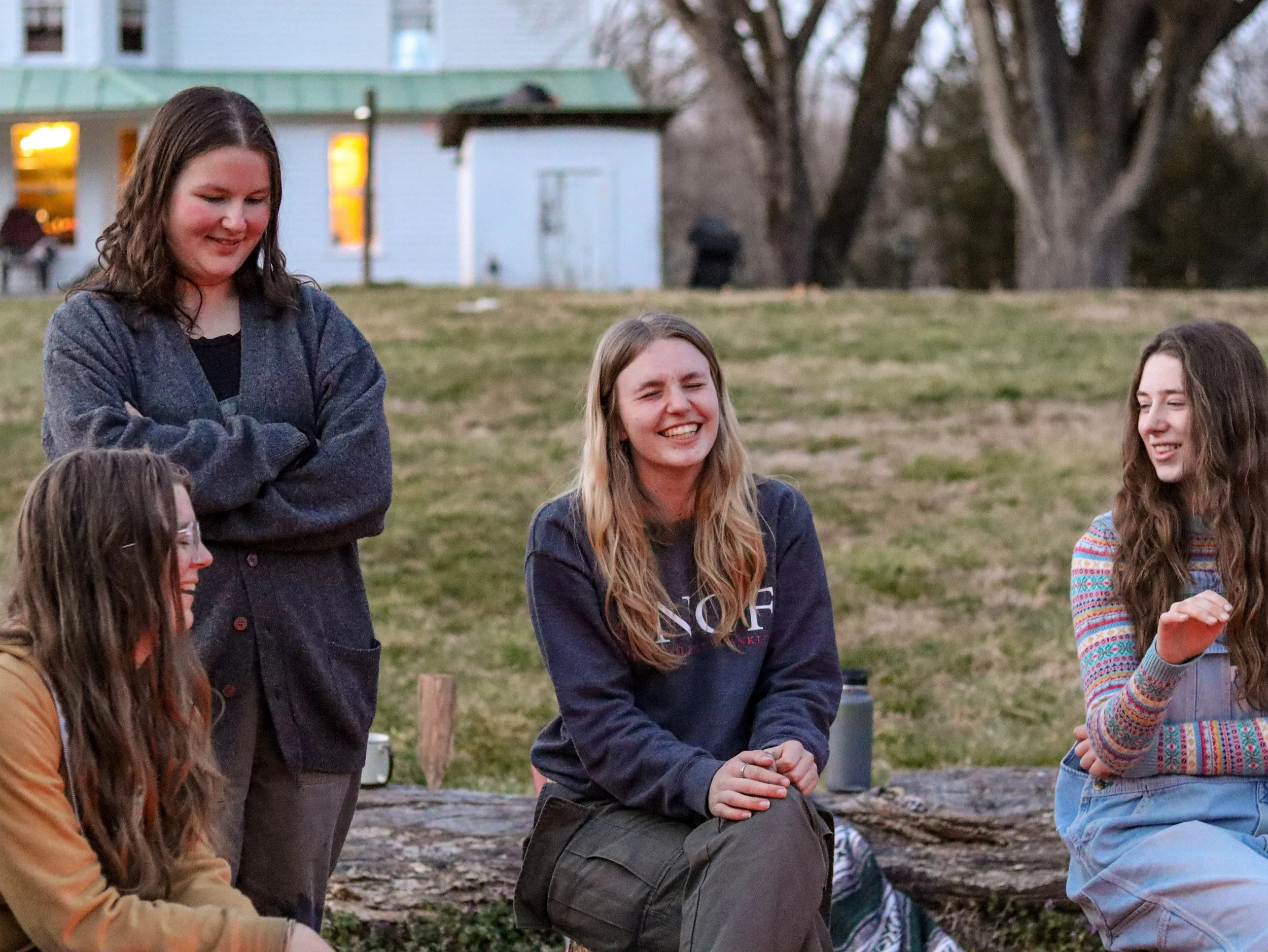Group of young women gathered outdoors in a park-like setting, sitting and standing, engaging in conversation and smiling. In the background are trees, a white house, and a green roof structure, with a warm evening light.