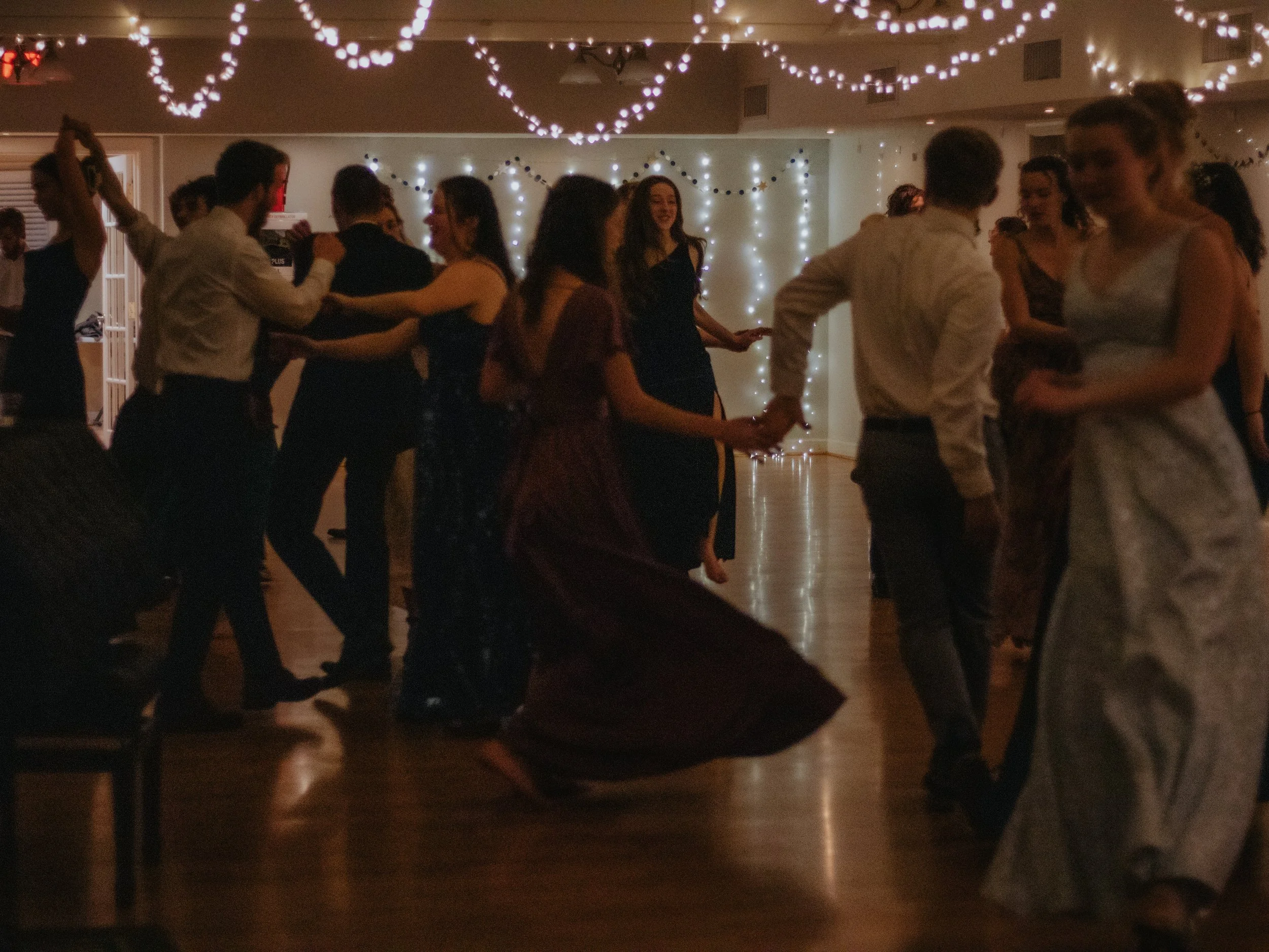 People dancing at a celebration with string lights and wall decorations in the background.