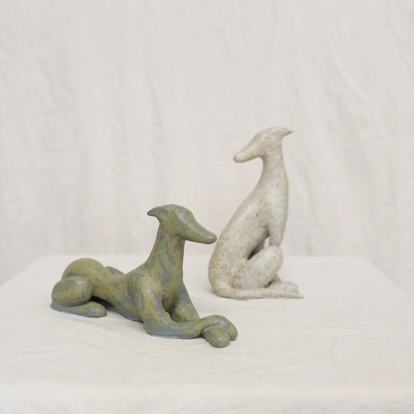 Two abstract, ceramic dog sculptures, one lying down and the other sitting, against a plain background.