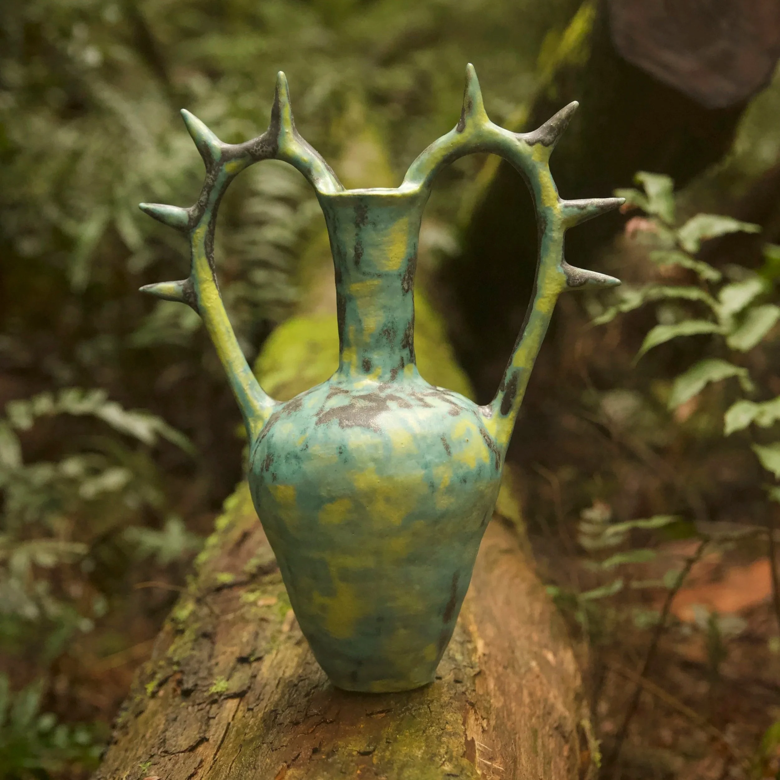 A ceramic vase with a heart-shaped opening and protruding spikes on each side, set outdoors on a tree trunk with a blurred natural background.