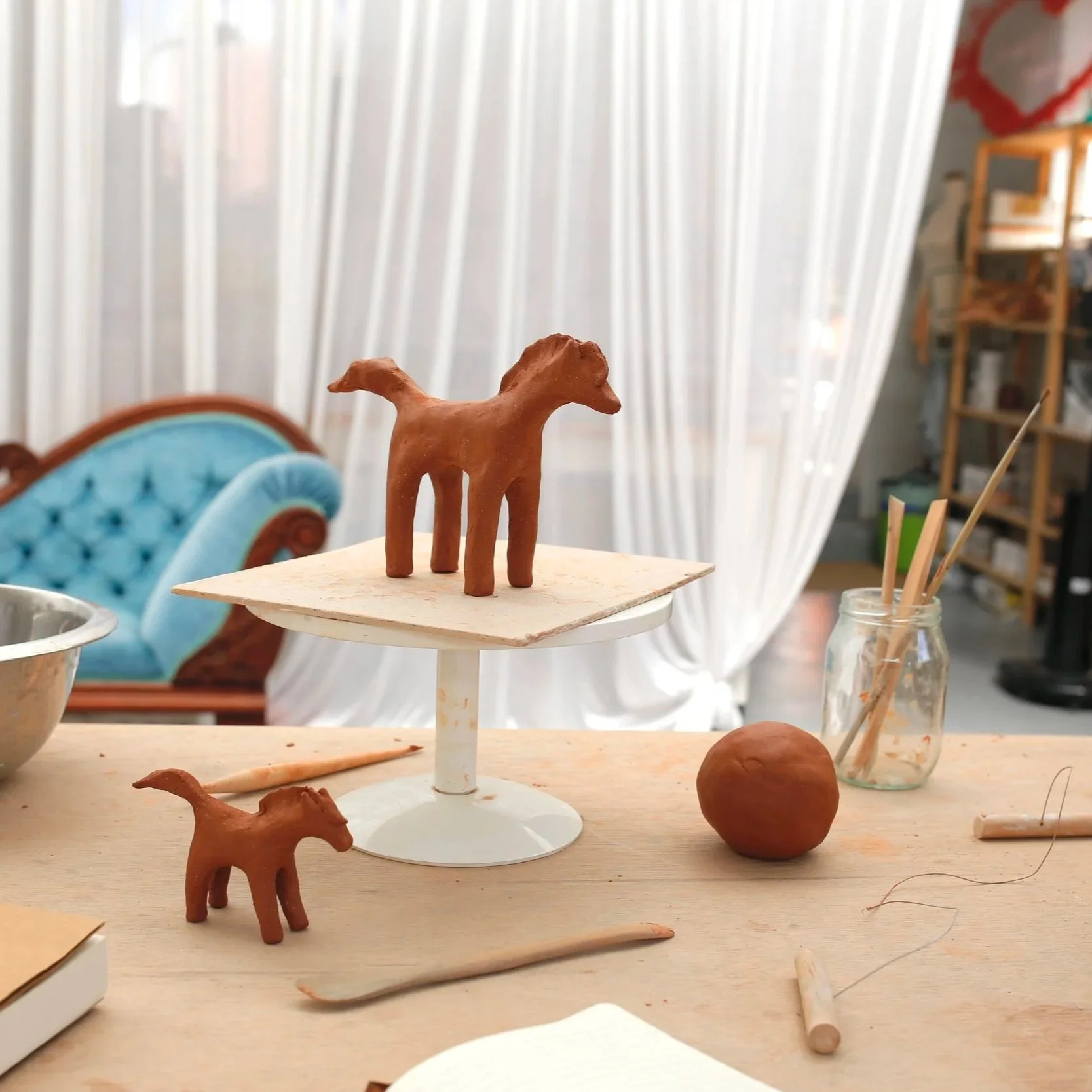 Clay horse sculptures on display, with one on a cake stand and another on the table, surrounded by pottery tools and materials in an art studio.