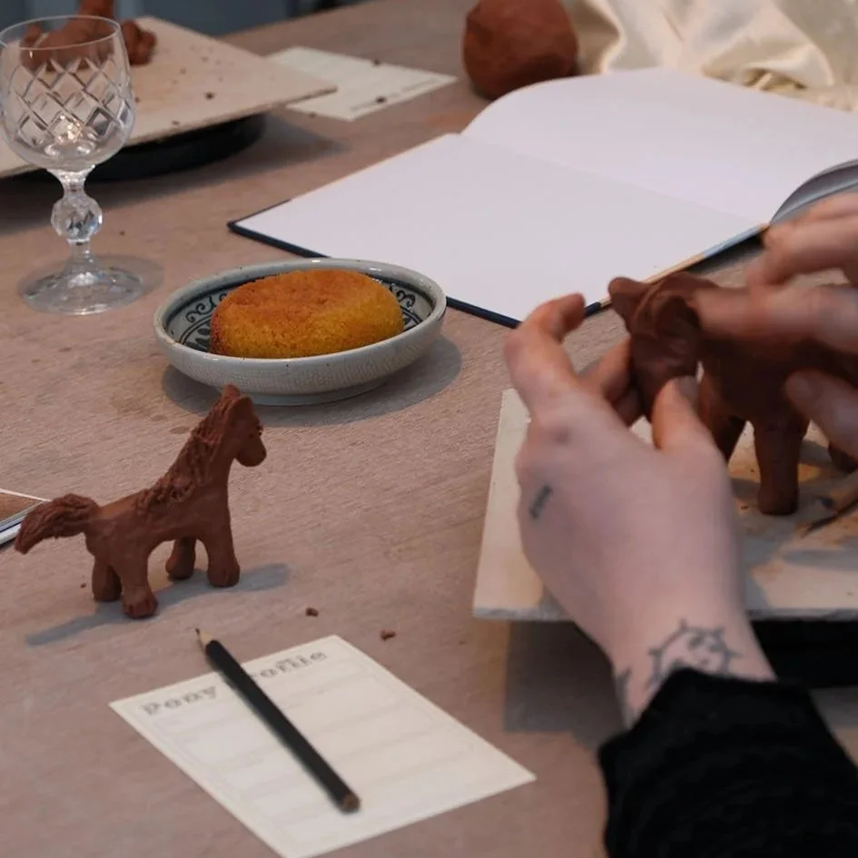 A person with tattoos on their hand making a clay horse sculpture on a wooden table. Other items on the table include a small horse figurine, a notepad, a pen, a dish with a round brown snack, an empty wine glass, and a notebook.
