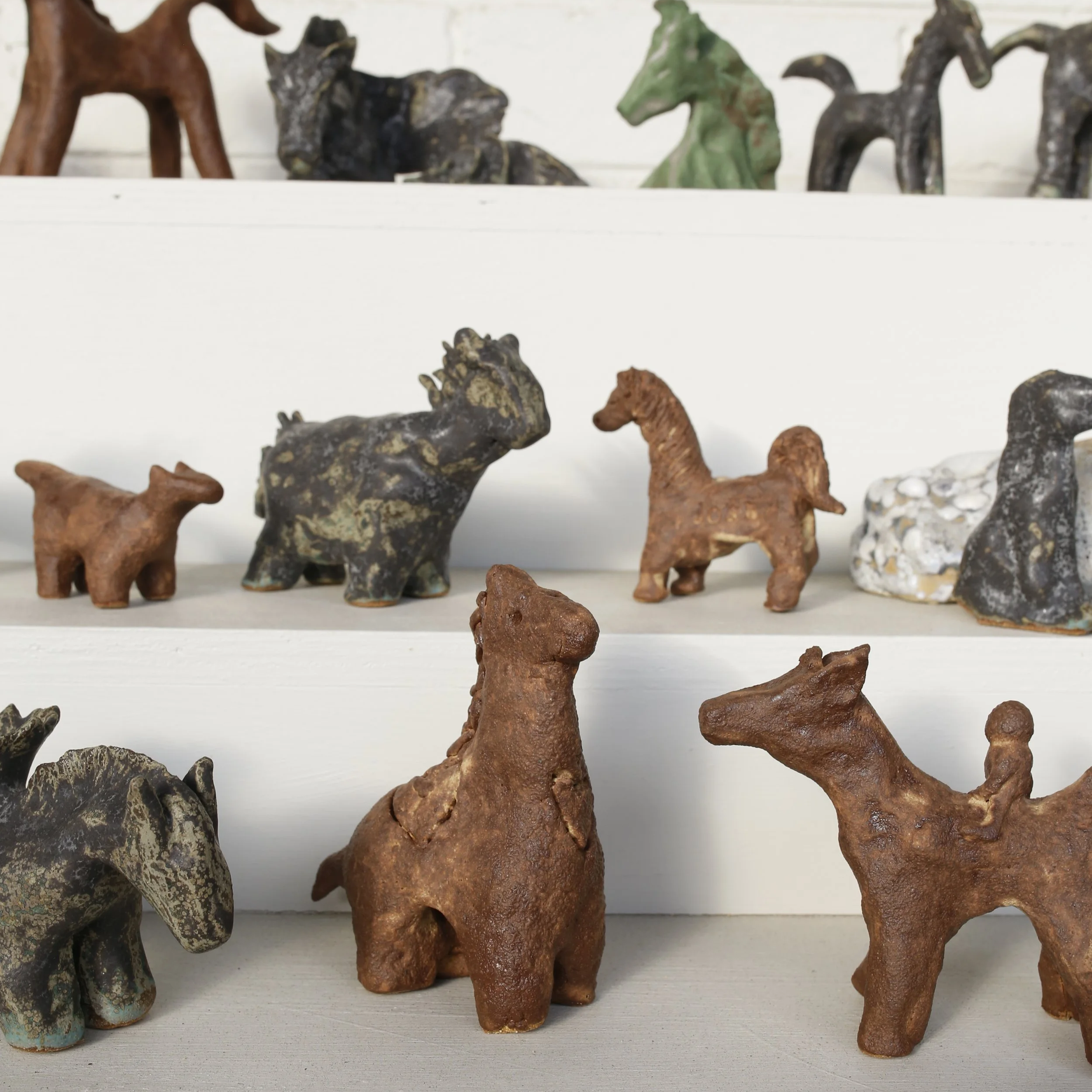 Collection of small, painted ceramic animal figurines on white shelves, including horses, a dog, and other animals.
